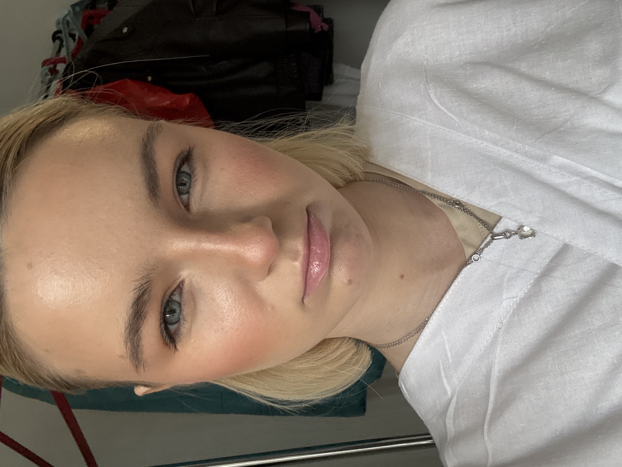 A close-up, indoor portrait shows a fair-skinned young woman with light blonde hair. She has prominent cheekbones with blush and pale blue eyes. She is wearing a white linen shirt and a delicate silver necklace. The background includes what appears to be a messy bed or storage area with dark-colored clothing items. The image is rotated, giving the impression the subject is lying down. There is nothing to indicate the location is in Saint Petersburg, Russia.