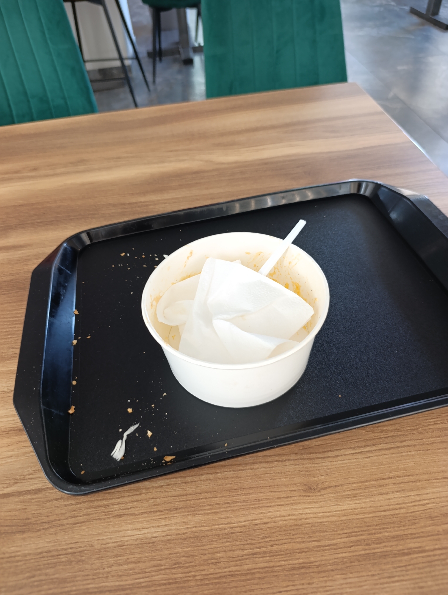 A black tray sits on a wooden table, containing a white paper cup with some food remnants and a few crumpled napkins. A white plastic straw protrudes from the cup. Crumbs are scattered on the tray and table. In the background, green chairs are visible. The image appears to be taken indoors, possibly in a cafe or restaurant setting. There is no information about the location in the city of Dubăsari town council, Moldova.