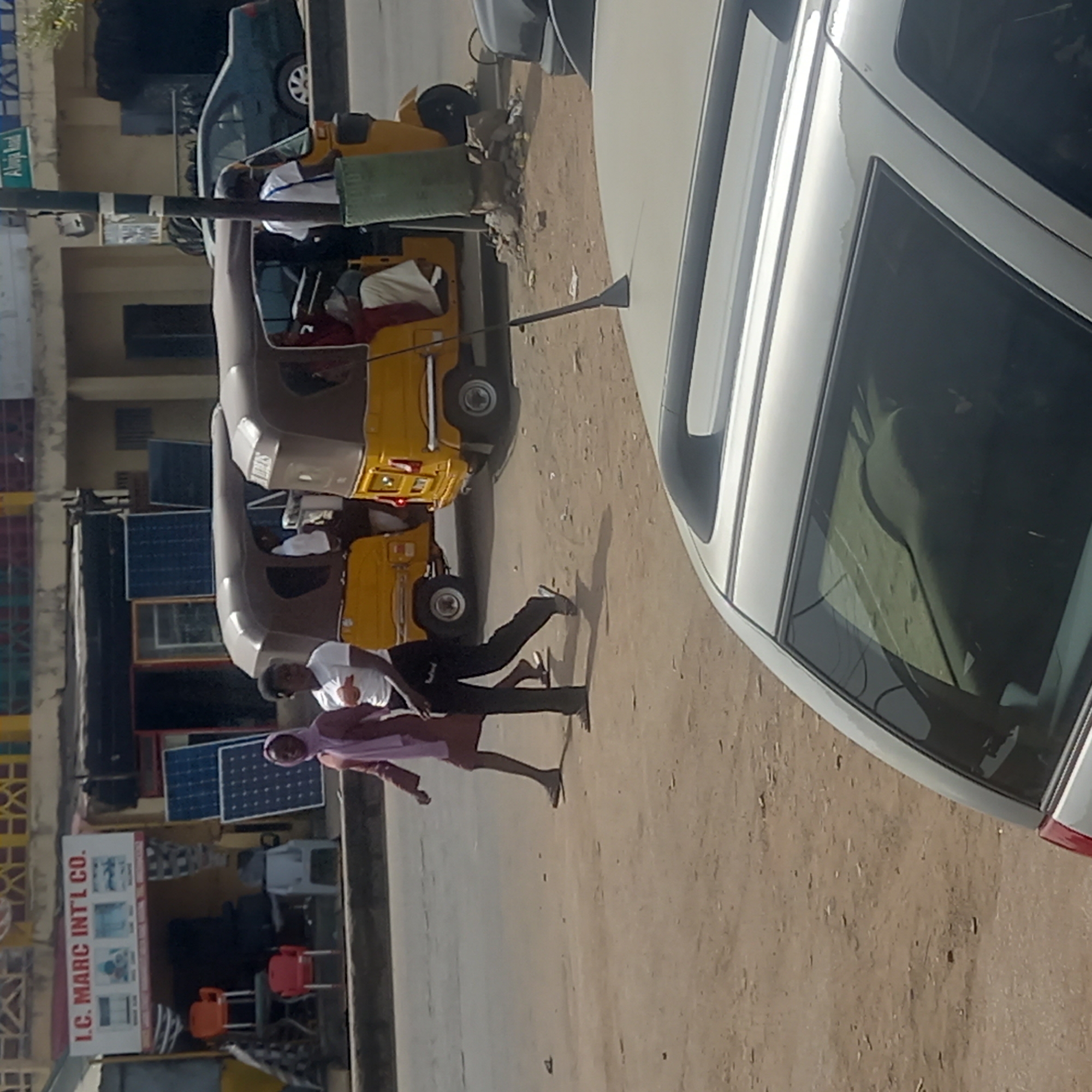 This image captures a vibrant street scene in Dadi Riba, Nigeria, under bright, clear daylight conditions. The time of day appears to be around midday or early afternoon, indicated by the distinct, relatively short shadows cast by people and objects on the dusty ground.
In the foreground and mid-ground, two individuals are seen walking on the unpaved roadside. One person wears a white t-shirt and dark trousers, while the other is dressed in a light purple hooded garment and lighter-colored pants. Both are barefoot or wearing sandals.
Several vehicles populate the scene. A light-colored car (possibly white or silver) is partially visible on the far right. Two Keke NAPEPs (tricycle taxis), a common form of public transport in Nigeria, are present on the road: one in a brown/tan color and another yellow one further back. Passengers in white shirts are visible inside both Keke NAPEPs. In the background, a dark-colored car is also visible near a street pole.
On the left side of the road, various commercial activities are evident. A shop displays a large white sign that reads "I.C. MARC INT'L CO." along with images of appliances like a refrigerator, suggesting it might be an electronics or home goods store. Outside this shop, red and orange chairs are placed on display. Several blue solar panels are mounted vertically against a building, hinting at a local energy solution. A green street sign in the background, partially visible, clearly states "Abuja Road," providing a significant location cue within Nigeria.
The setting depicts a typical busy street in a Nigerian town or city, characterized by a mix of pedestrian activity, local transportation, and roadside businesses.