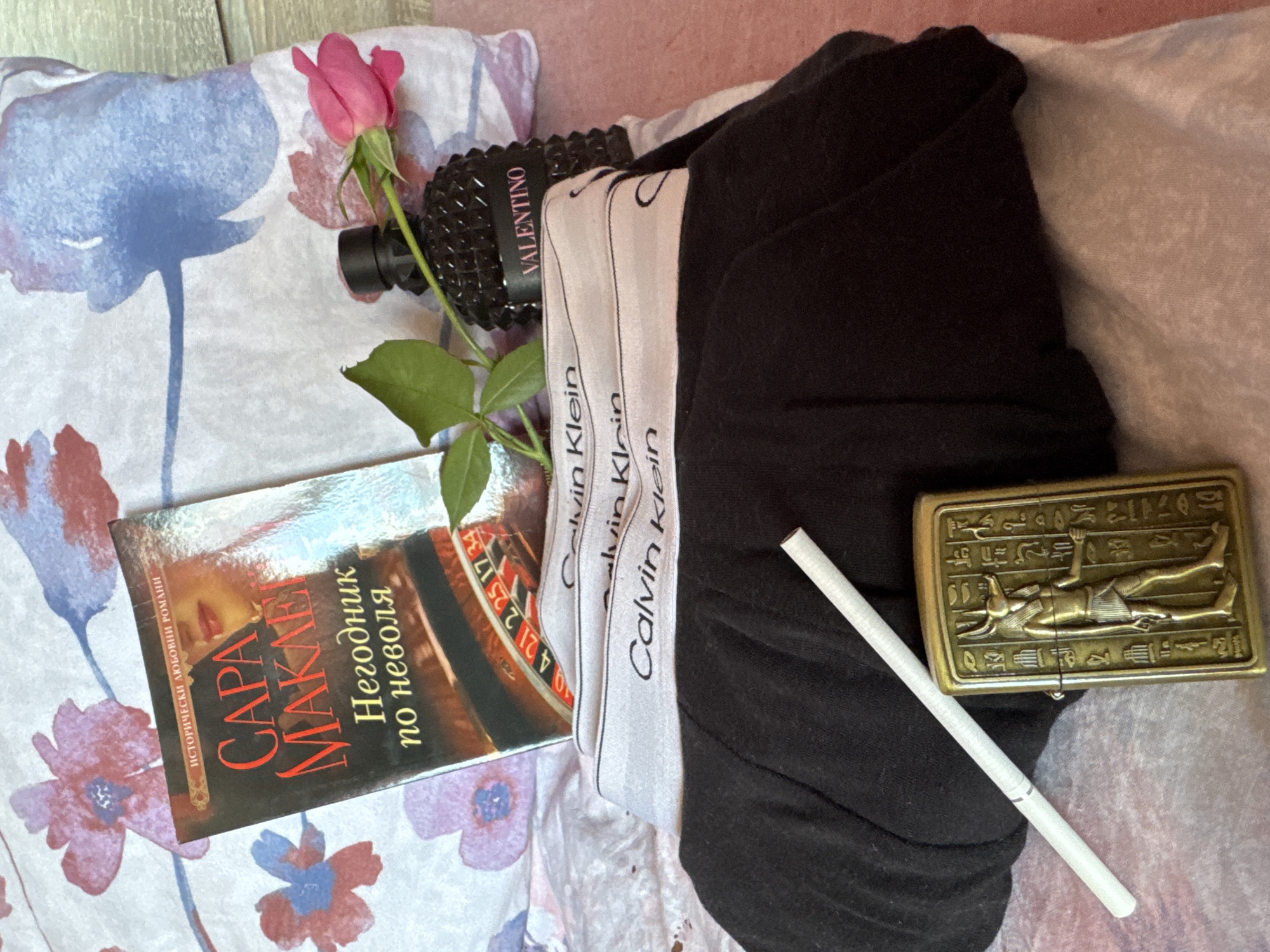 The image is a still life composition of personal items arranged on a floral patterned fabric. A pink rose with green leaves lies next to a black Valentino bottle. A book with the title "CAPA MAKAEI Негодник по неволя" is visible with a partial image of a roulette wheel on its cover. Three Calvin Klein branded waistbands are stacked next to the book and Valentino bottle, partially obscured by a pile of black clothing. A gold-colored, rectangular item depicting an Egyptian figure and hieroglyphs rests next to a white cigarette. The items are arranged on a floral-patterned sheet on a pink surface. There is no indication this image was taken in Dolna Oryahovitsa, Bulgaria.