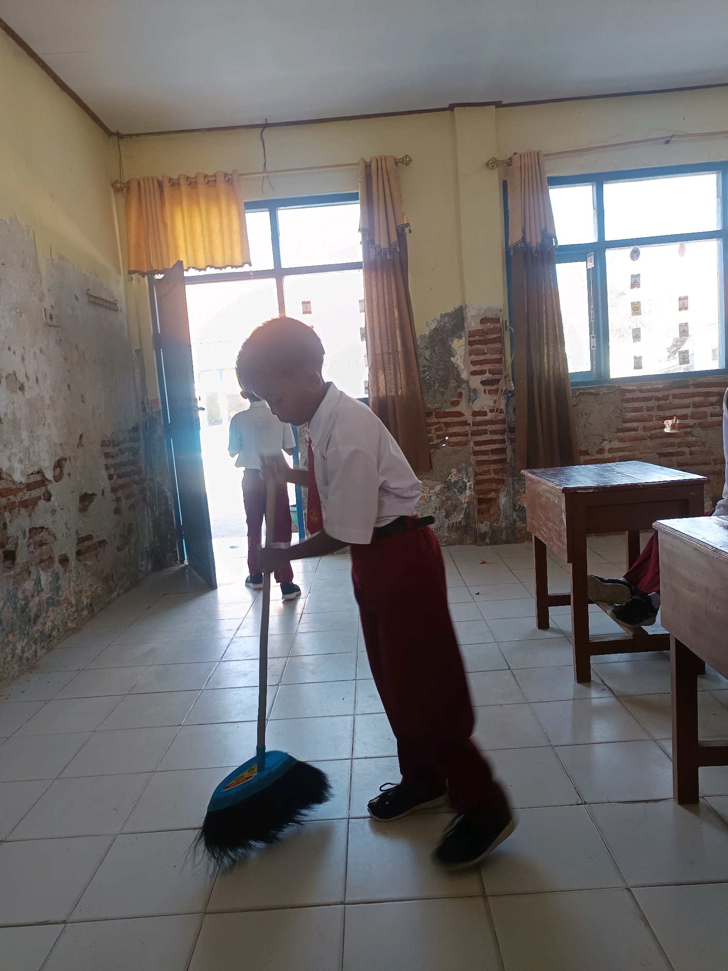 In Labuhan, Indonesia, a young student in a white shirt and maroon pants is sweeping a tiled floor with a blue broom. Another student, also in uniform, stands in the background near an open doorway. The room appears to be a classroom, with desks visible and peeling paint and exposed brick on the walls. Sunlight streams in through windows covered by tan curtains.