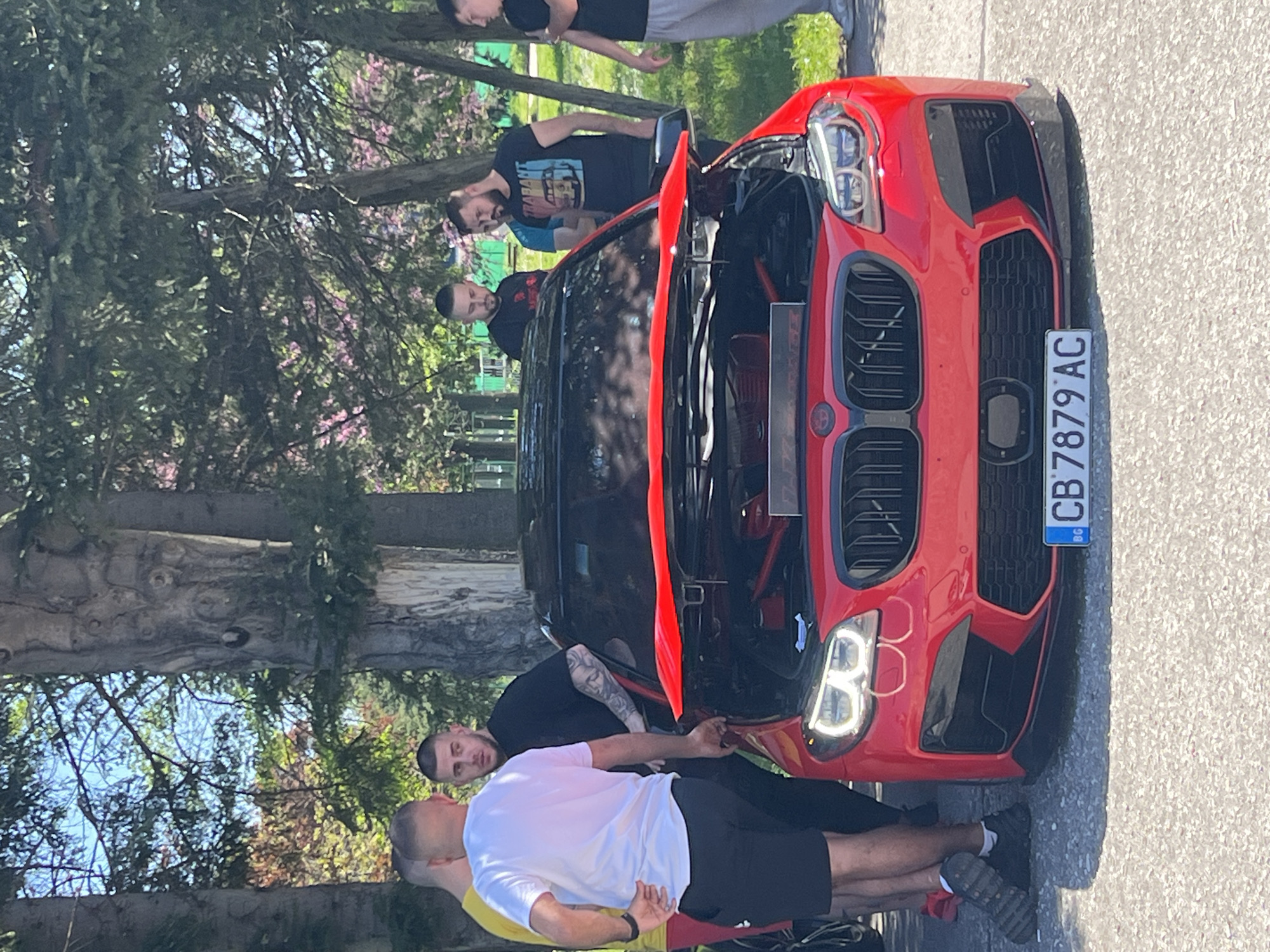 A red BMW car with an open hood is parked on a paved surface. Several men are gathered around the vehicle. The license plate is visible and reads "CB 7879 AC". The location is potentially in Plovdiv, Bulgaria, based on the "CB" license plate code, which is typical for the city. Trees are present in the background, with some flowering branches visible. The day appears sunny. Some men are wearing casual clothing, such as t-shirts and shorts. There appear to be a few spectators possibly observing the car.
