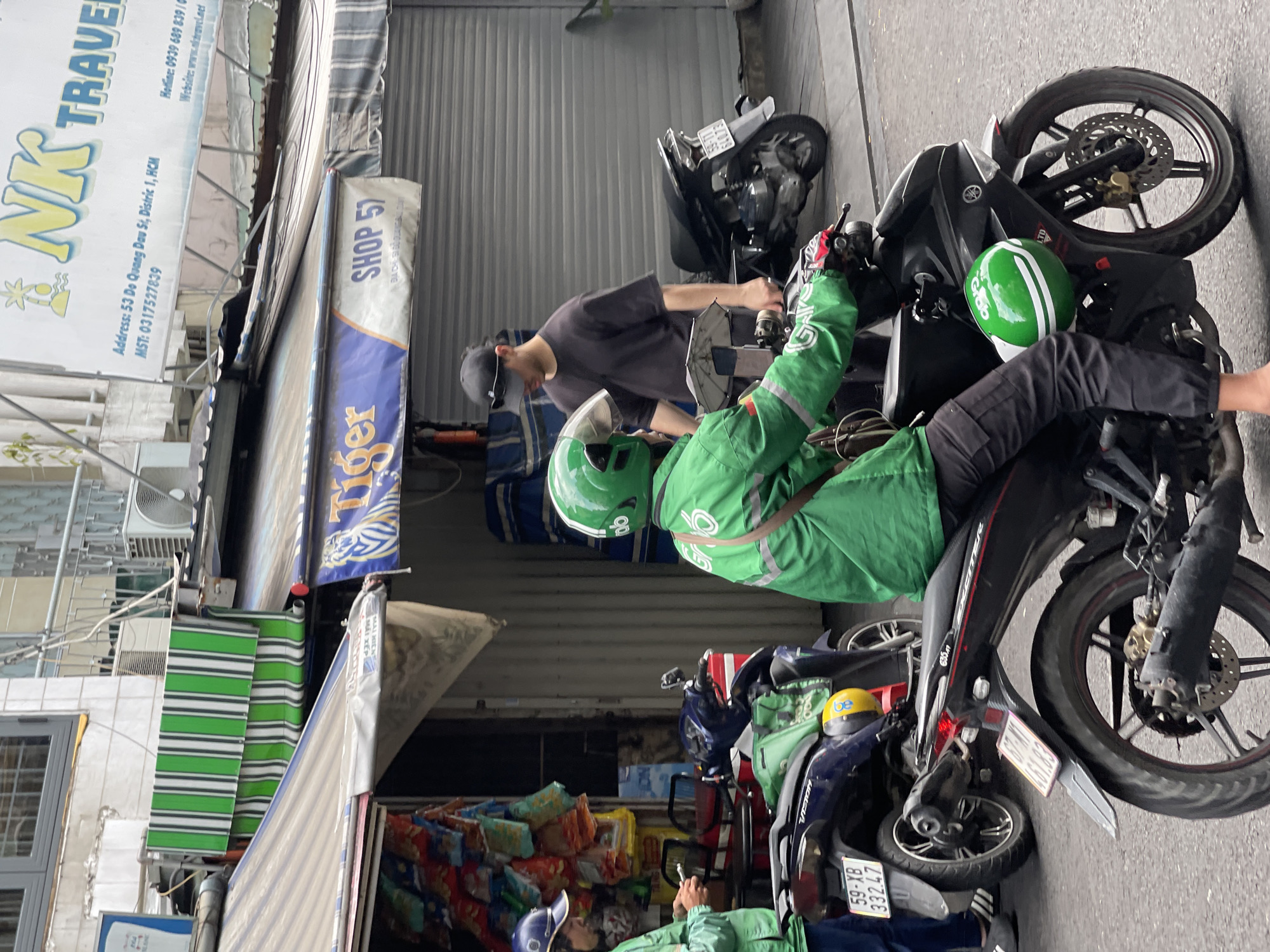 The image depicts a street scene in Thủ Đức, Vietnam, with several individuals and objects of interest. Two individuals are visible wearing green uniforms and helmets, likely delivery drivers. They are near multiple parked motorbikes, a common mode of transportation in Vietnam. The storefronts feature Vietnamese signage, including a "Tiger" banner, the name "NK TRAVEL" and address "53 Do Quang Dau St, Distric 1, HCM." Some awnings are striped green and white. The shop is labeled "Shop 57". License plates on the motorbikes are visible. One motorbike has a license plate of "59-XB 332.47." Another plate is "461.83."