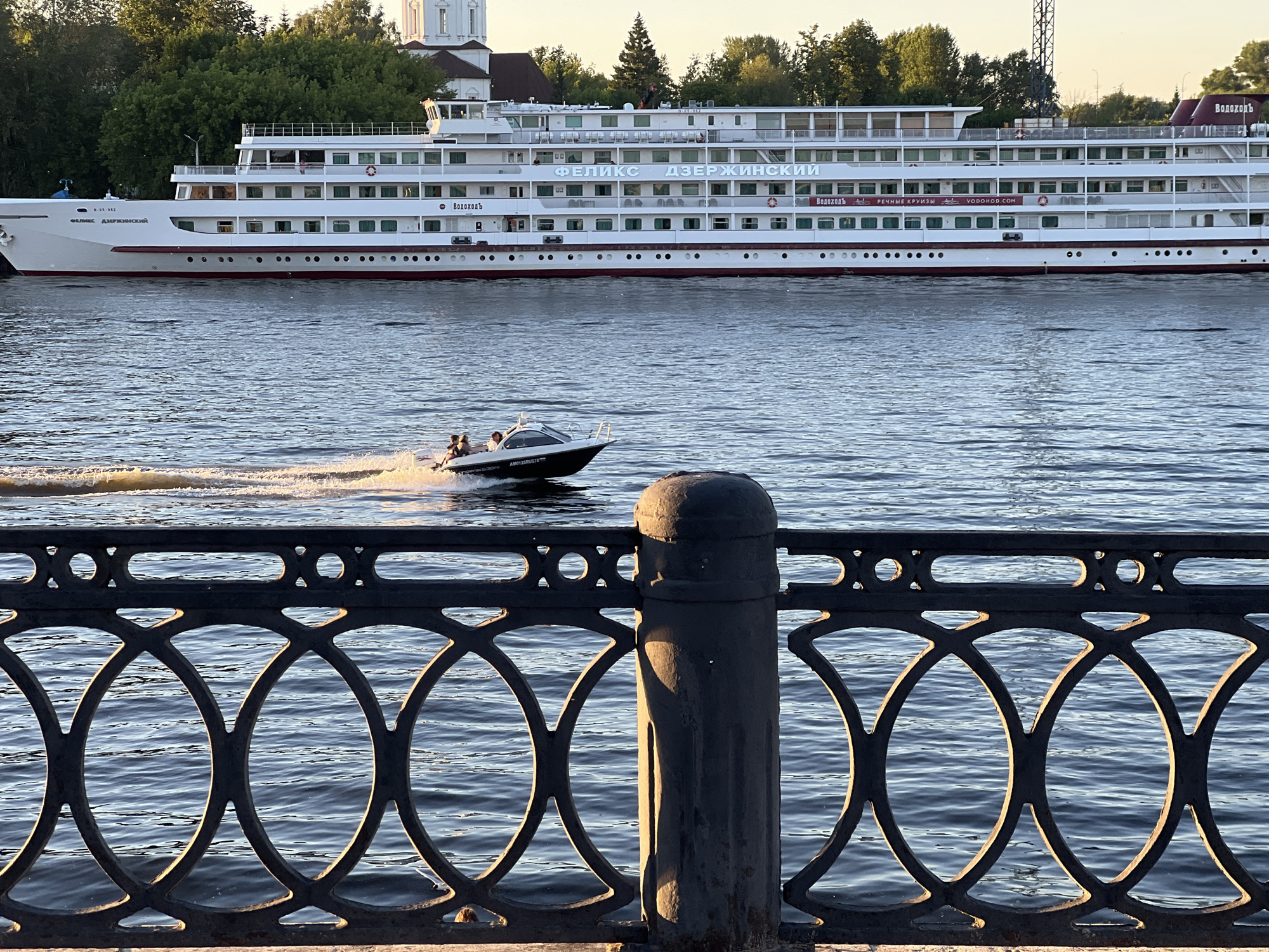 A large white passenger ship, named "FELIKS DZHERZHINSKIY" in Cyrillic, is moored on the Volga River in Tver, Russia. The ship has multiple decks with numerous windows and red accents along the hull. In the foreground, a black metal railing with decorative circular patterns separates the viewer from the river. A small motorboat with three passengers is speeding across the water, creating a wake. The sun is setting, casting a warm glow on the scene and illuminating the water and the railing. Trees and buildings, including a white church-like structure, are visible on the far bank of the river. The sky is a soft orange and yellow, indicating the end of the day.