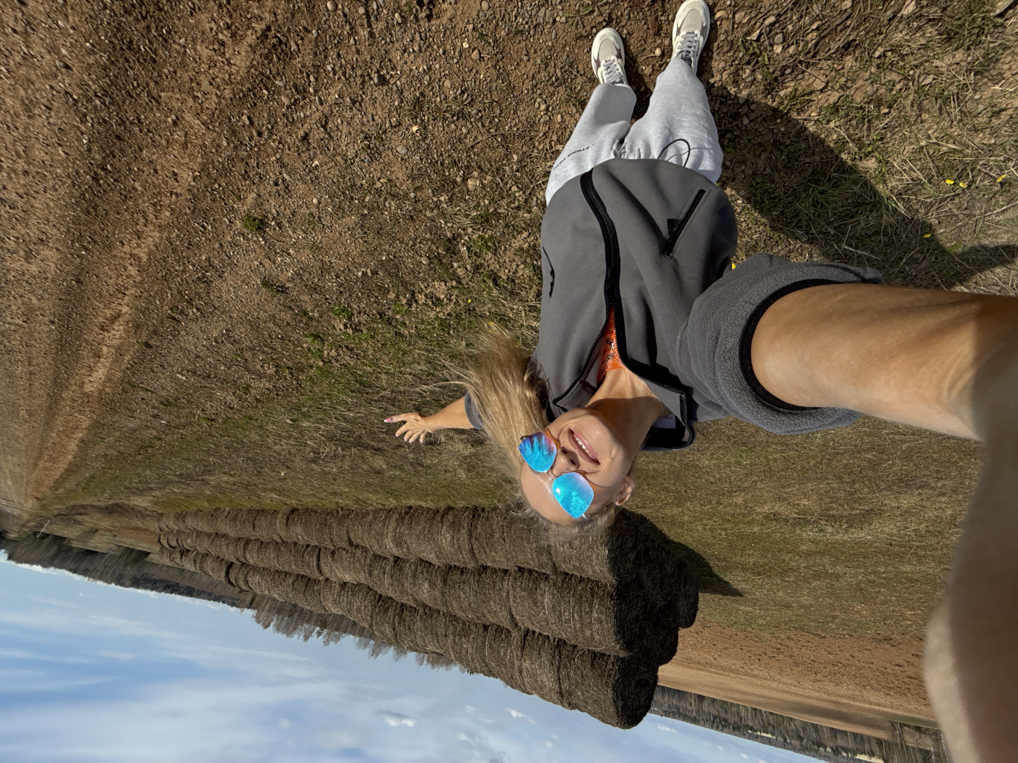 A high-angle selfie captures a smiling blonde woman lying on dry, gravelly ground, looking upwards towards the camera held by her outstretched right arm. She is wearing blue reflective sunglasses, a dark grey zip-up jacket over a lighter top, light grey sweatpants with "STRAND UP" faintly visible on the left leg, and light-colored sneakers. Her body is oriented horizontally across the frame, with her left arm casually extended to the side, suggesting a relaxed pose in an open field.
The ground she lies on is a mixture of earth, small stones, and sparse patches of dry grass. Behind her, a long, neatly stacked row of dark brown round hay bales stretches horizontally across the midground of the scene, indicating an agricultural setting. Beyond the hay bales, a flat, light brown field extends to the horizon, where a dense line of trees forms a dark silhouette against the sky. The upper portion of the image reveals a clear, bright blue sky with scattered white clouds, characteristic of a sunny day, likely midday or afternoon.
The image conveys a sense of outdoor leisure and joy in a rural landscape, consistent with the location context of Малая Уса, Russia.