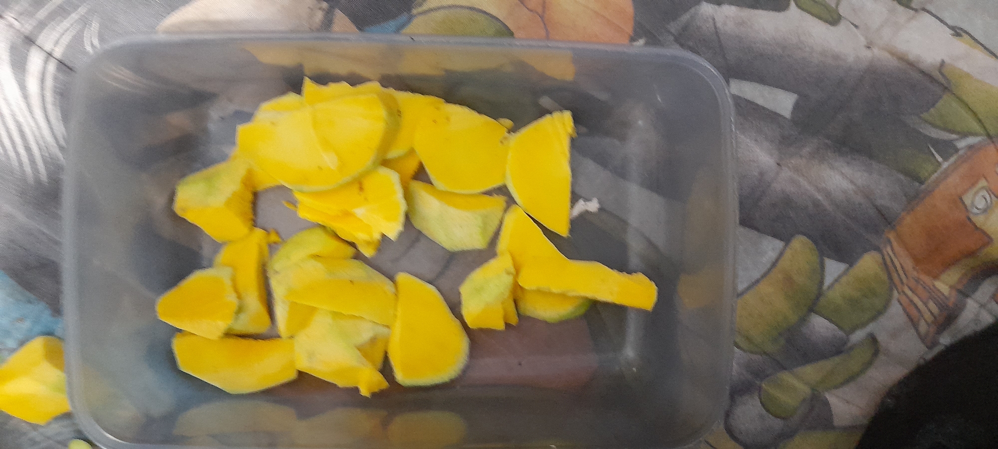 The image shows a grey plastic container filled with slices of ripe mango. The container is sitting on a patterned surface with shades of grey, green, and brown. The image was taken in Buketteukuh, Indonesia.