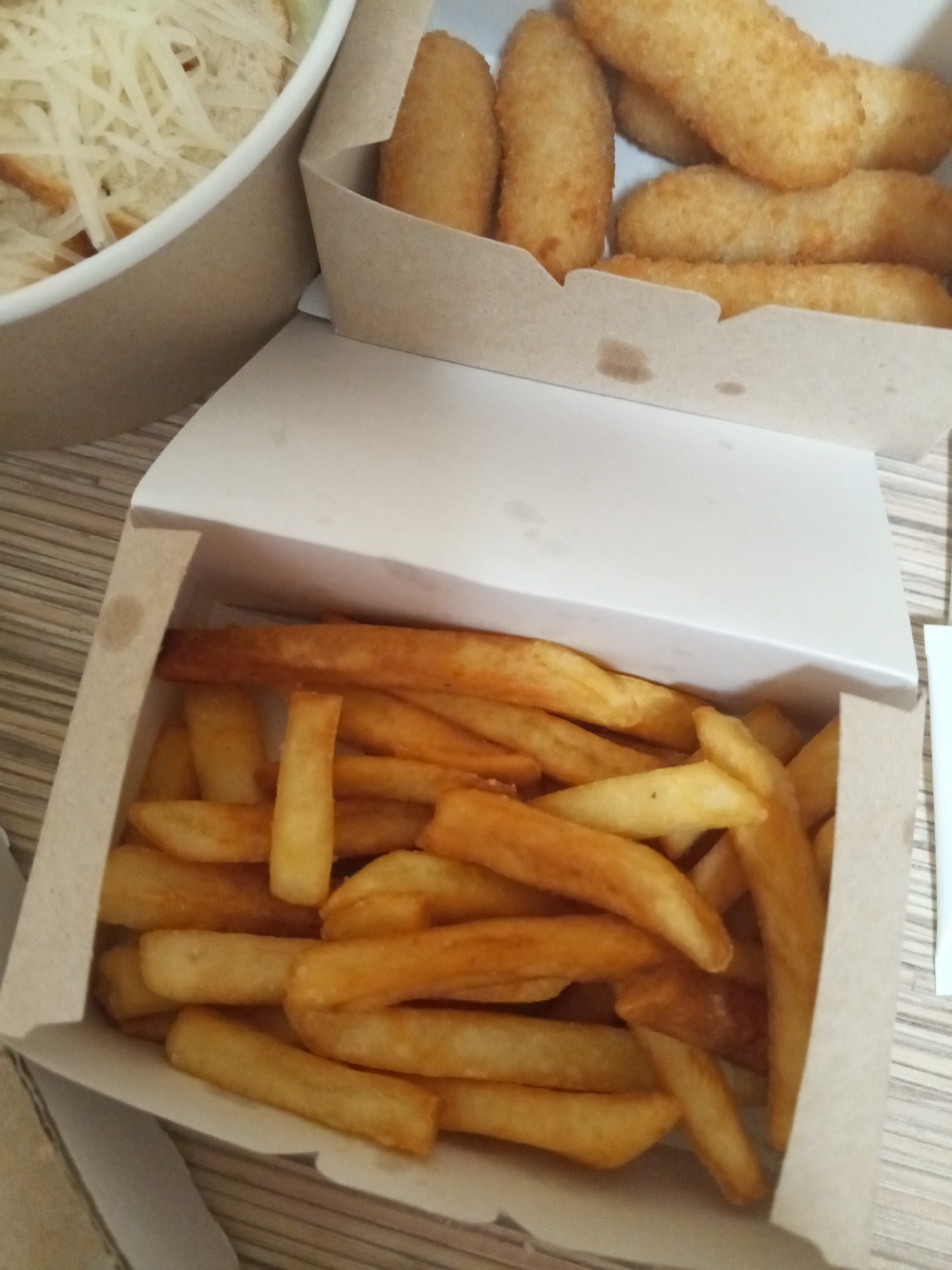 Two cardboard boxes of food are placed on a light brown wooden surface. The box in the foreground contains a large portion of golden-brown french fries. The box in the background is filled with several golden-brown fried items, possibly chicken tenders or mozzarella sticks, and is partially obscured by the lid of the foreground box. To the left of the boxes, a white bowl is visible, filled with a shredded white substance, likely cheese, on top of bread or croutons. The overall scene suggests a meal or take-out food. There are no people or animals present in the image. The lighting is somewhat dim, with shadows cast from the boxes onto the surface. The location is Chistopol, Russia.