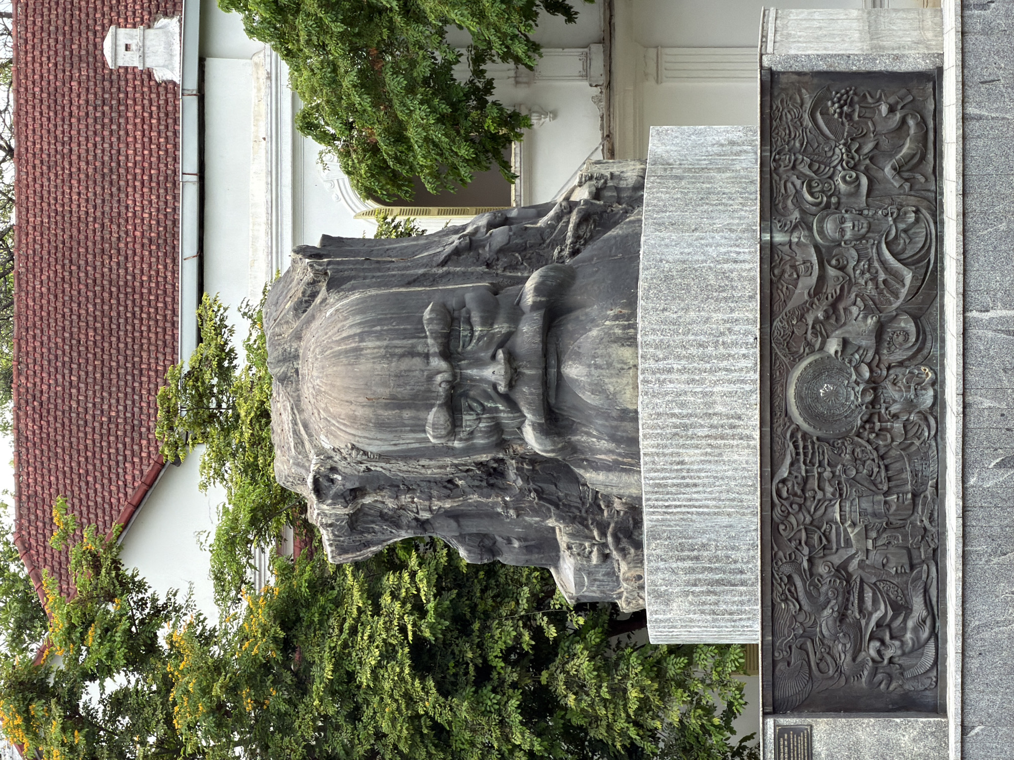 The image shows a large, dark stone sculpture of a man's head, likely a historical figure. The sculpture is mounted on a tiered, grey granite pedestal. Below the head sculpture, a large, dark bronze bas-relief panel depicts various scenes and figures, including mythical creatures like dragons, and what appear to be human faces and traditional architectural elements. The entire monument is set against a backdrop of a white building with a red tiled roof, partially obscured by lush green trees with some yellow flowers. The setting suggests an outdoor public space, likely in a city given the architectural style and monument. Based on the provided context, this monument is located in Phú Xuân Ward, Vietnam.