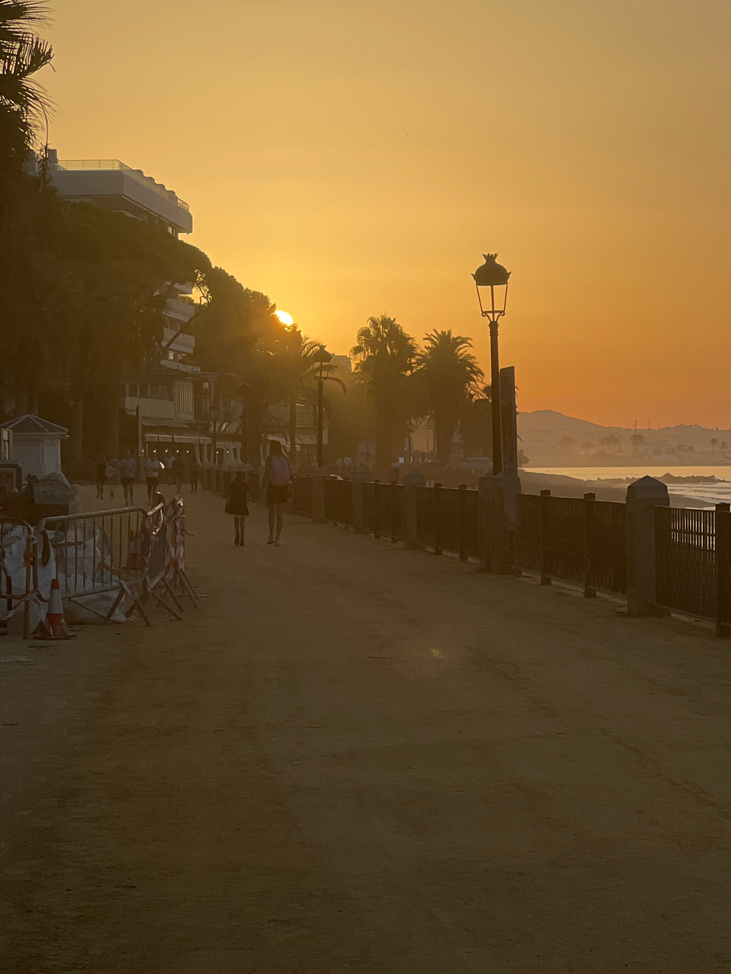 A sunset on the promenade in Marbella, Spain. The sun is partially visible, casting a warm orange glow over the sky and sea. A paved walkway runs along the coast, with a dark metal fence separating it from the beach. Several people are walking on the promenade, some with children. Palm trees and buildings line the left side of the walkway. A lamppost stands tall near the fence. Construction barriers are visible on the left side of the path. The scene evokes a sense of tranquility and end-of-day calm.