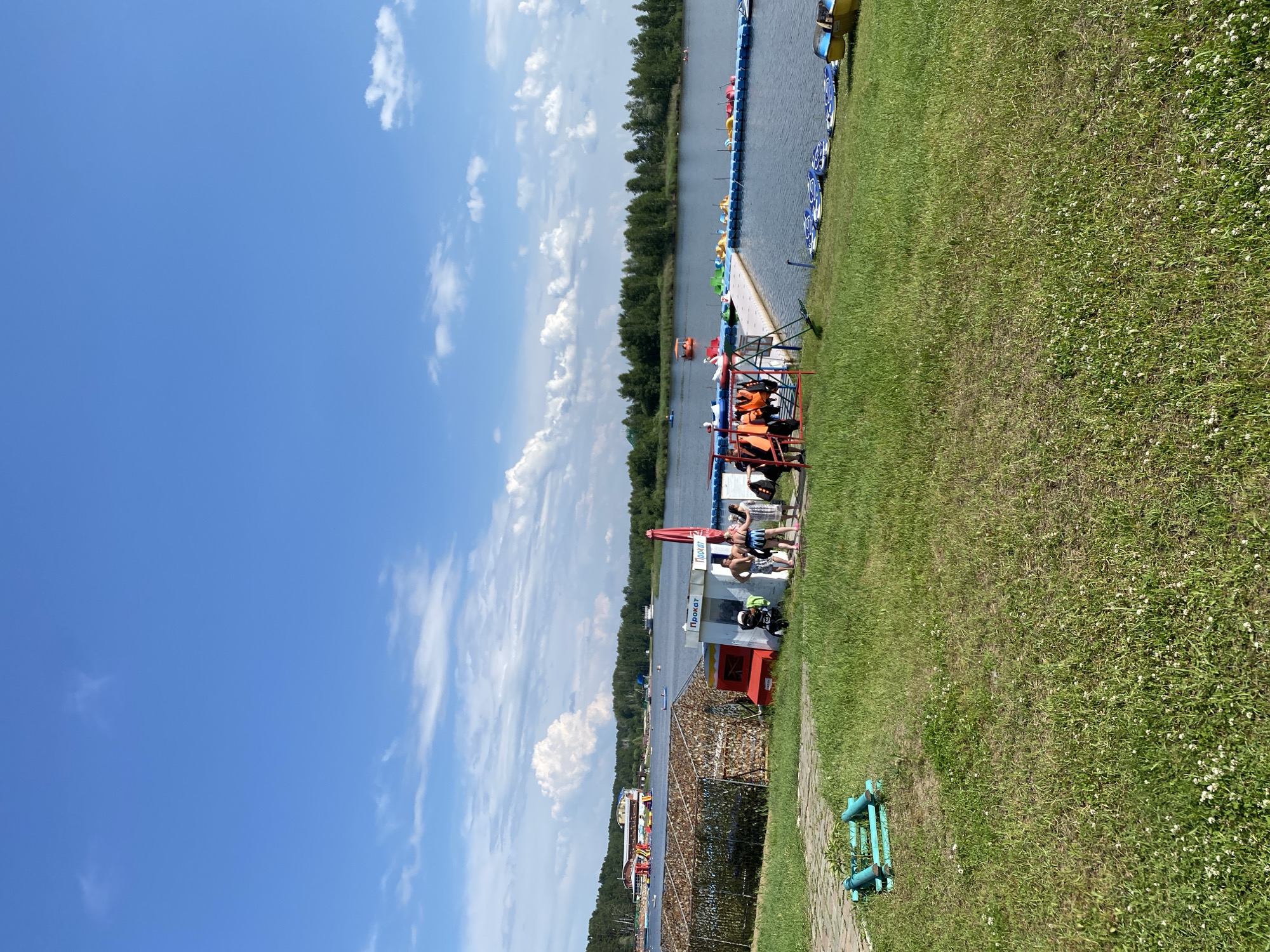 The image depicts an outdoor recreational area by a lake on a sunny day with scattered clouds. In the foreground, a grassy area with some small white flowers is visible, along with a blue tubular structure. A paved path leads to a small building with "Прокат" (Rental) signs. Several people are standing near this building, some appearing to be rental staff. Adjacent to the building is a rack holding several orange life vests.
Further into the scene, a floating dock extends into the lake, lined with colorful paddle boats and other watercraft. Across the lake, a treeline forms the horizon, and a multi-story building with red and yellow structures, possibly a resort or water park, is visible in the distance. Several boats are scattered on the lake, and more trees are visible on the far bank. The overall impression is a vibrant summer day at a lake resort.