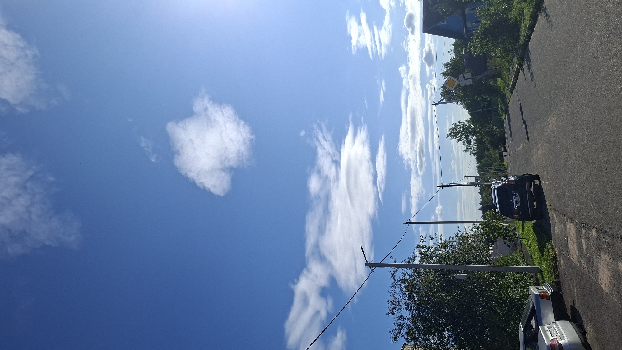 This is a vertical, eye-level shot of a paved road in a rural or suburban area, likely in Myadzyel, Belarus, on a bright, sunny day with a clear blue sky dotted with fluffy white clouds. The road curves slightly to the right and upwards in the distance. On the left side of the road, parked parallel to the curb, are two cars: a silver sedan and a darker sedan or hatchback. Further down the road on the left, a black SUV is parked on the side of the road. There are trees and houses visible on either side of the road. On the left, there is a dense green tree and a portion of a white fence. On the right side of the road, there is a blue house with a white trim and a pitched roof, with green bushes and trees surrounding it. A yellow diamond-shaped road sign with a black border and a black symbol indicating a curve to the right is mounted on a pole near the blue house. Telephone poles and wires are visible throughout the scene. In the far distance, a few small figures can be seen on the road, possibly walking or cycling.