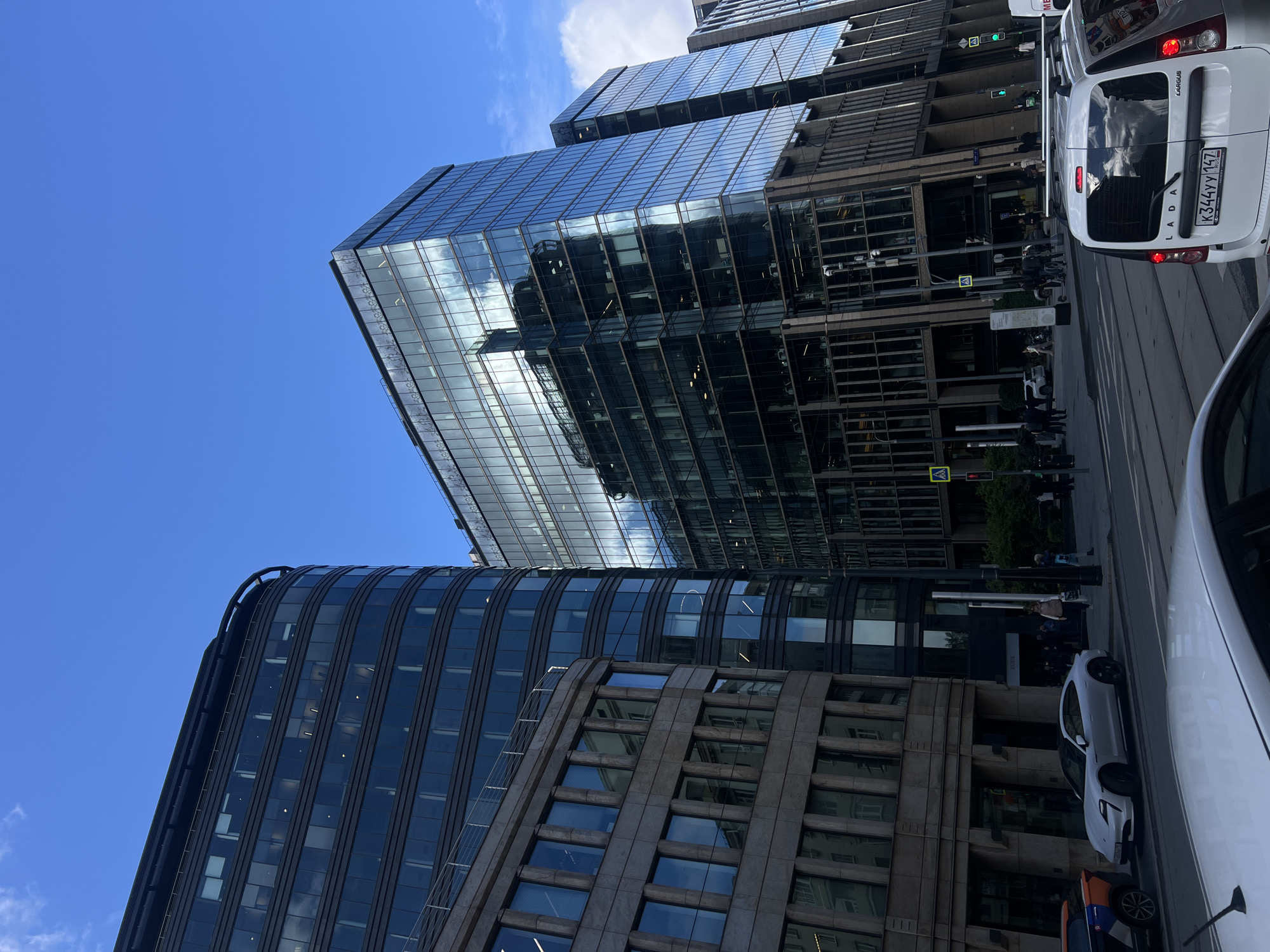 The image depicts a street scene in Moscow, Russia, distinguished by modern architecture, vehicular traffic, and pedestrian activity. The architectural style is contemporary, featuring glass and steel structures, typical of commercial districts. The weather is bright and sunny, with a clear blue sky. There are several cars visible on the road, including a white Lada Largus with a license plate number K344YY147, indicating Russian registration. Pedestrians can be seen on the sidewalks. The presence of street signs and traffic signals indicates a regulated urban environment.
