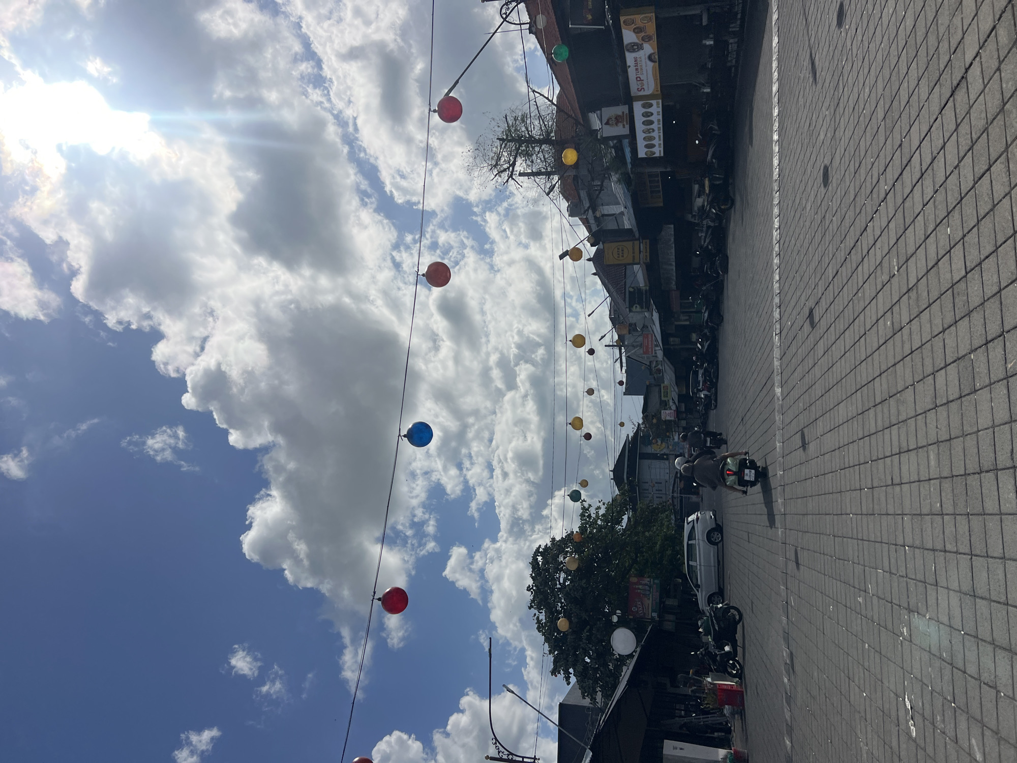 This image depicts a street scene in Kuta, Indonesia, characterized by a bright, sunny day with scattered clouds. The street is paved with interlocking gray tiles, and a line of shops with dark awnings lines the right side of the road. Overhead, strings of colorful spherical lanterns in red, blue, yellow, and green are strung across the street, adding a festive atmosphere. On the left side of the street, more shops and trees are visible, along with parked motorcycles. A white SUV is parked in the middle distance, and several people are on motorcycles, with one person riding a scooter away from the camera. The sun is high in the sky, casting shadows and creating a bright glare in the upper right portion of the image. The overall impression is of a lively, bustling commercial area on a clear day.