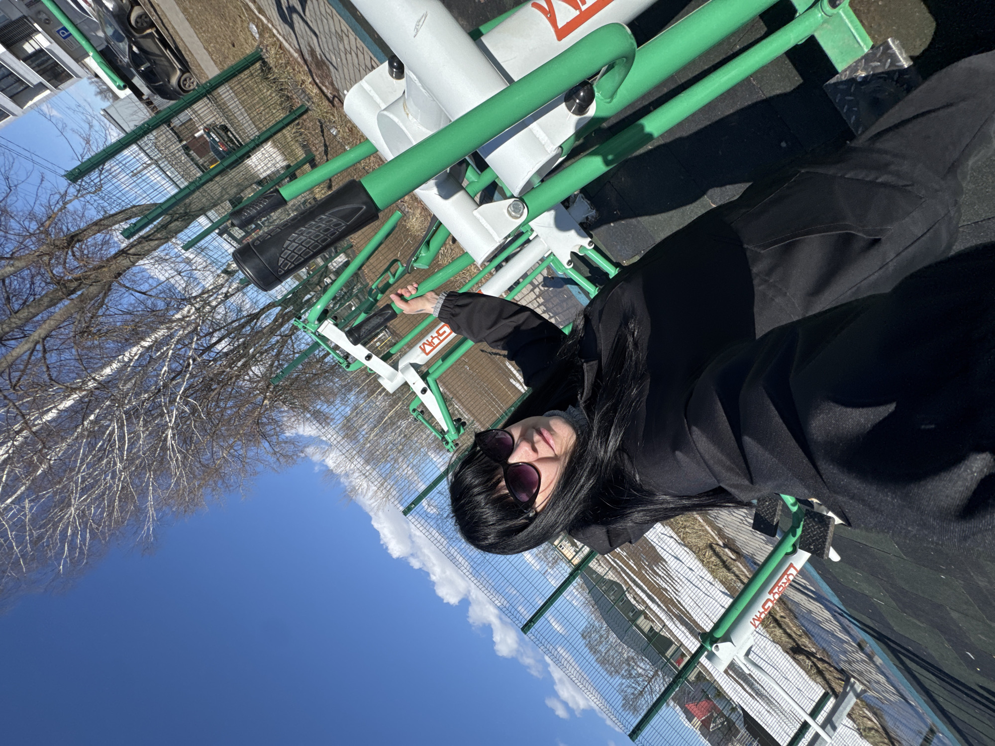 A woman with long black hair and sunglasses is in an outdoor gym in Zav'yalovo, Russia. She is dressed in black and appears to be operating the exercise equipment, which is painted green and white. The sky is blue with some clouds, and bare trees are visible in the background, suggesting it is early spring. A wire fence surrounds the gym area. Other workout stations are also present.
