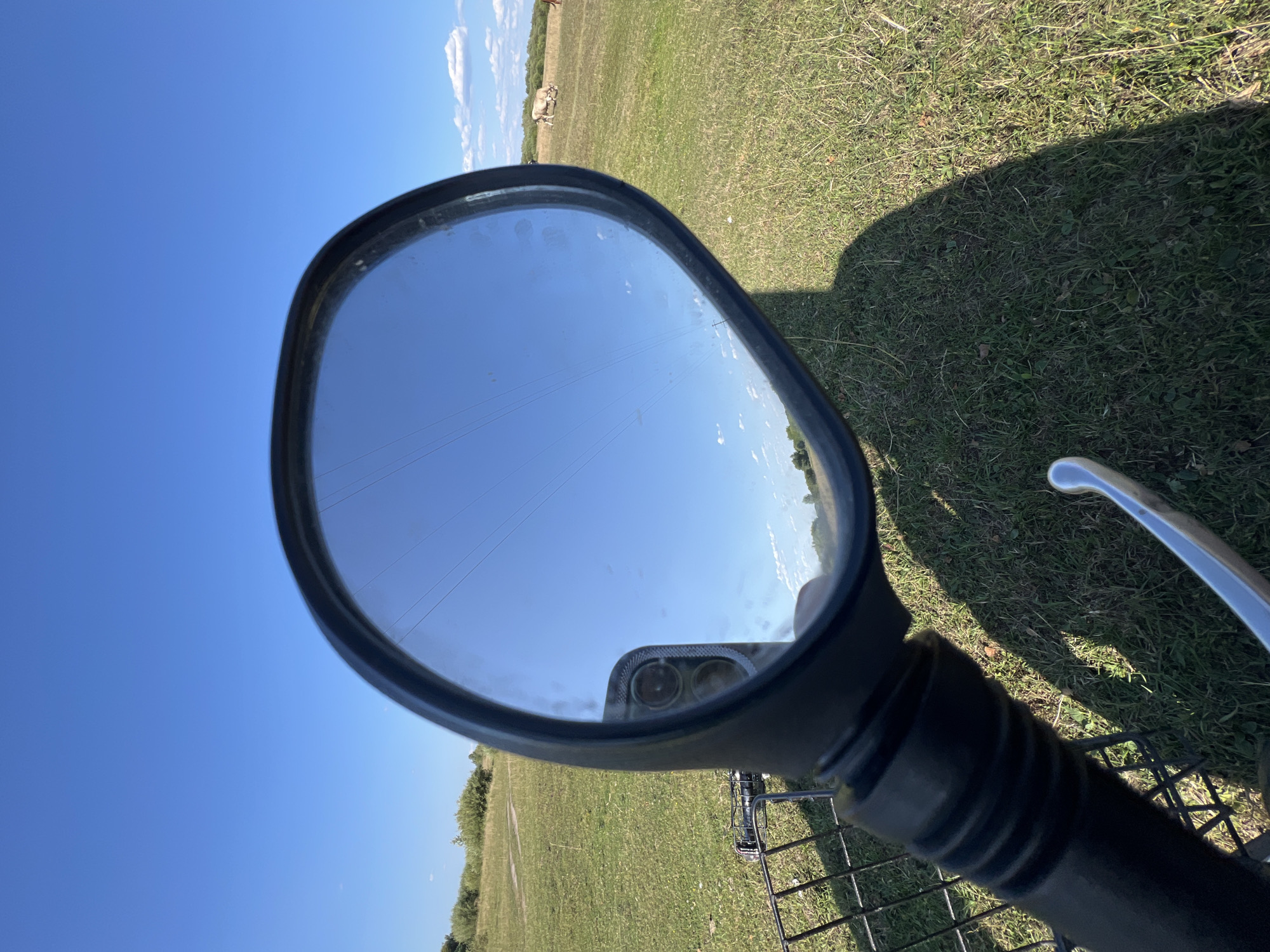 This image, captured in the vicinity of Sobishchytsi, Ukraine, depicts a close-up view of a rearview mirror on what appears to be a bicycle or motorcycle. The mirror is oval-shaped with a black rim and reflects a bright blue sky with thin, horizontal lines, possibly power lines. The foreground shows the textured black grip of the handlebar and a chrome brake lever. A dark shadow of the viewer is cast across the grassy ground, which is illuminated by bright sunlight. In the background, a green field stretches out under the sky, dotted with some bushes and trees on the horizon. Two light-colored cows are visible grazing in the field to the right. The overall scene suggests an outdoor environment on a clear day.