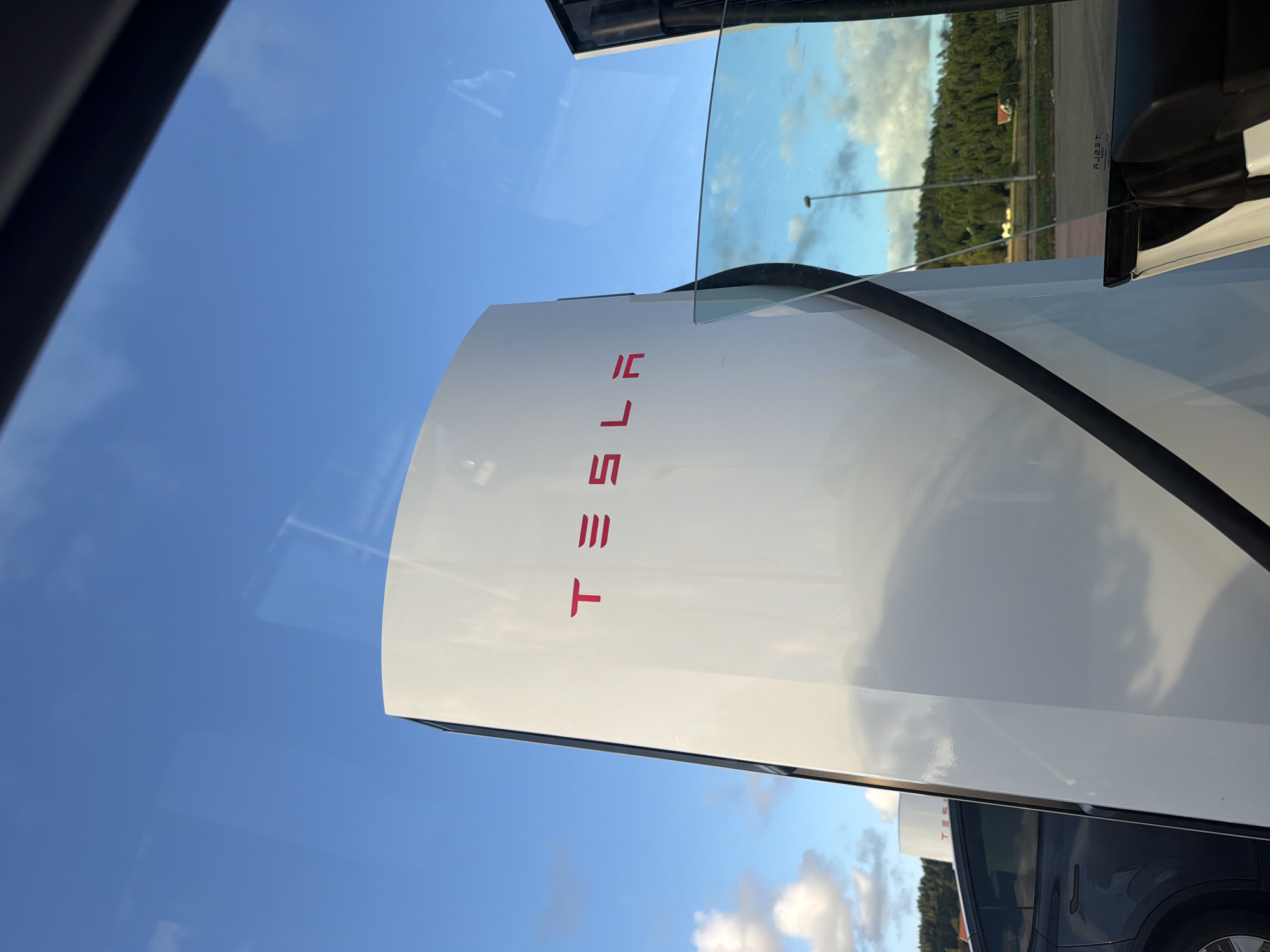 A Tesla Supercharger station is visible in the foreground under a blue sky with scattered clouds. The white, curved structure of the charging station features the prominent red Tesla logo. To the left, a dark-colored Tesla electric vehicle is parked. In the background, a tree-covered hillside is visible with a few buildings. The scene appears to be a public charging area. The image is taken from inside a vehicle, as suggested by reflections on a glass surface. The location is identified as Hogstorp, Sweden.