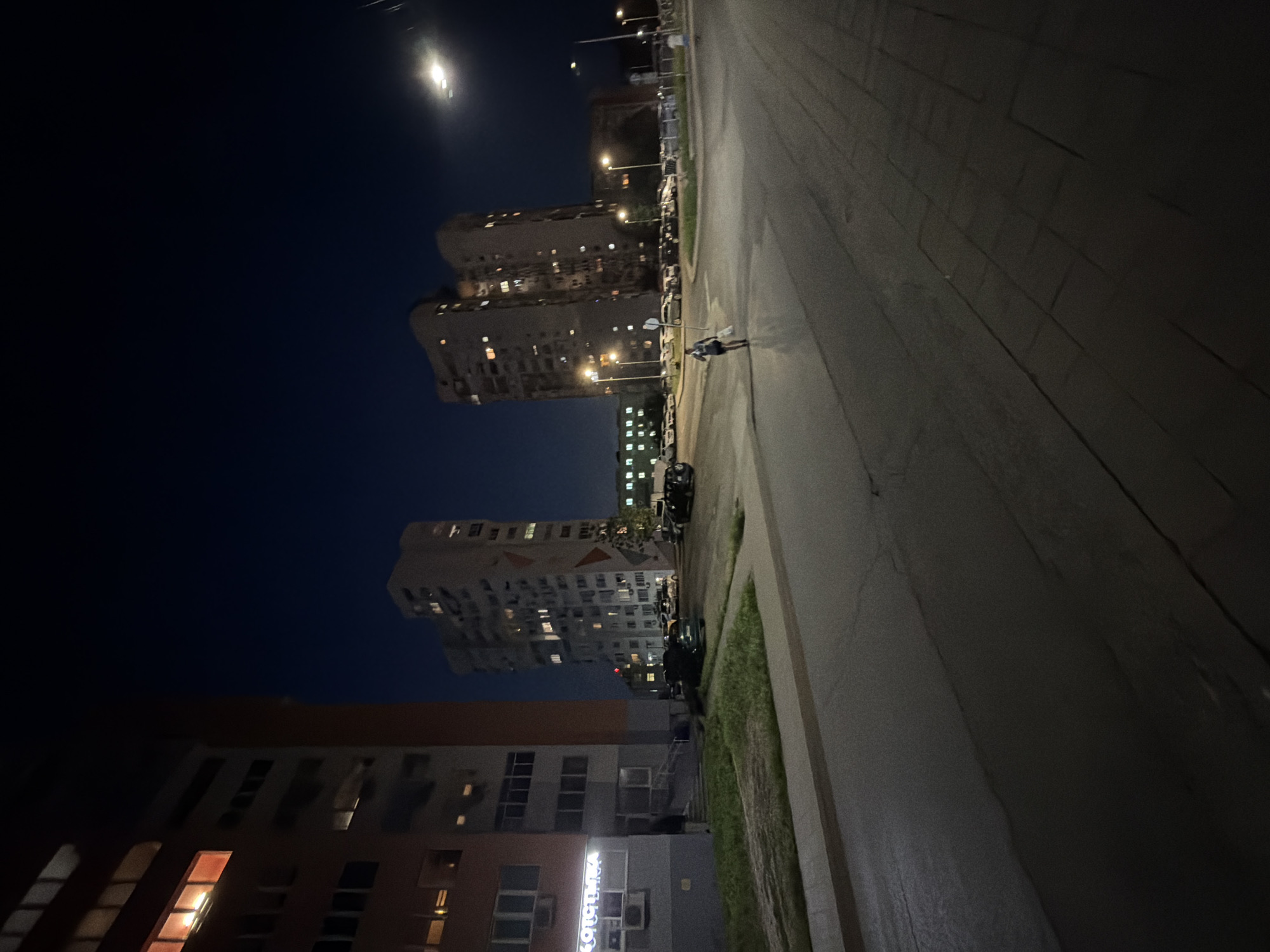 This nighttime image captures an urban residential street scene in Burgas, Bulgaria. The sky is dark, indicating a late hour, with artificial light sources illuminating the area.
On the left, a multi-story apartment building with a composite reddish-brown and light gray facade is partially visible. A ground-level commercial establishment features a lit sign in Cyrillic text, reading "КОПЧЕНЦА" (KOPCHENZA), and a pedestrian ramp leads to its entrance. An outdoor air conditioning unit is mounted on the building's facade. Several windows on this building are illuminated, with one on an upper floor exhibiting a prominent orange glow.
Towards the center and right, two prominent, light-colored multi-story residential apartment buildings stand tall, featuring sections with geometric patterns on their facades. Numerous windows within these structures are lit.
The foreground and mid-ground are dominated by an asphalt road displaying visible wet patches that reflect light. A grassy verge separates the road from a paved sidewalk on the left. On the right, a wider pavement, constructed from rectangular slabs, is discernible. Multiple streetlights are active, casting pools of light onto the road and surrounding areas. A bright, diffuse light source is visible high in the dark sky towards the upper right.
One individual, dressed in dark shorts and a t-shirt, walks on the road in the middle distance, moving towards the viewer but angled slightly left. This person is holding a white bag or object in their left hand. A second, indistinct figure is faintly visible walking away in the far distance on the right sidewalk.
Numerous parked vehicles, predominantly dark-colored sedans and SUVs, are visible in parking areas situated between the buildings and alongside the road.