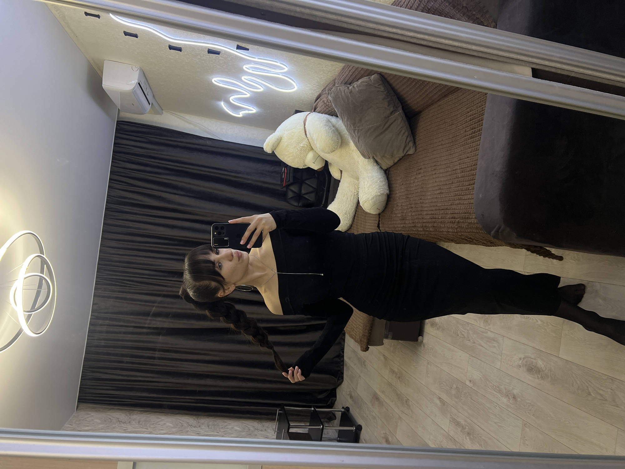 A person is taking a selfie in front of a mirror in an indoor environment. The individual is wearing a long-sleeved black dress. They have dark hair styled with bangs and a braid. The background includes a brown curtain, a brown couch with a brown pillow and a stuffed white bear. There's also a white neon light installation and a white air conditioner on the wall. The flooring appears to be wooden. The location cannot be verified as Votkinsk, Russia, without further information.