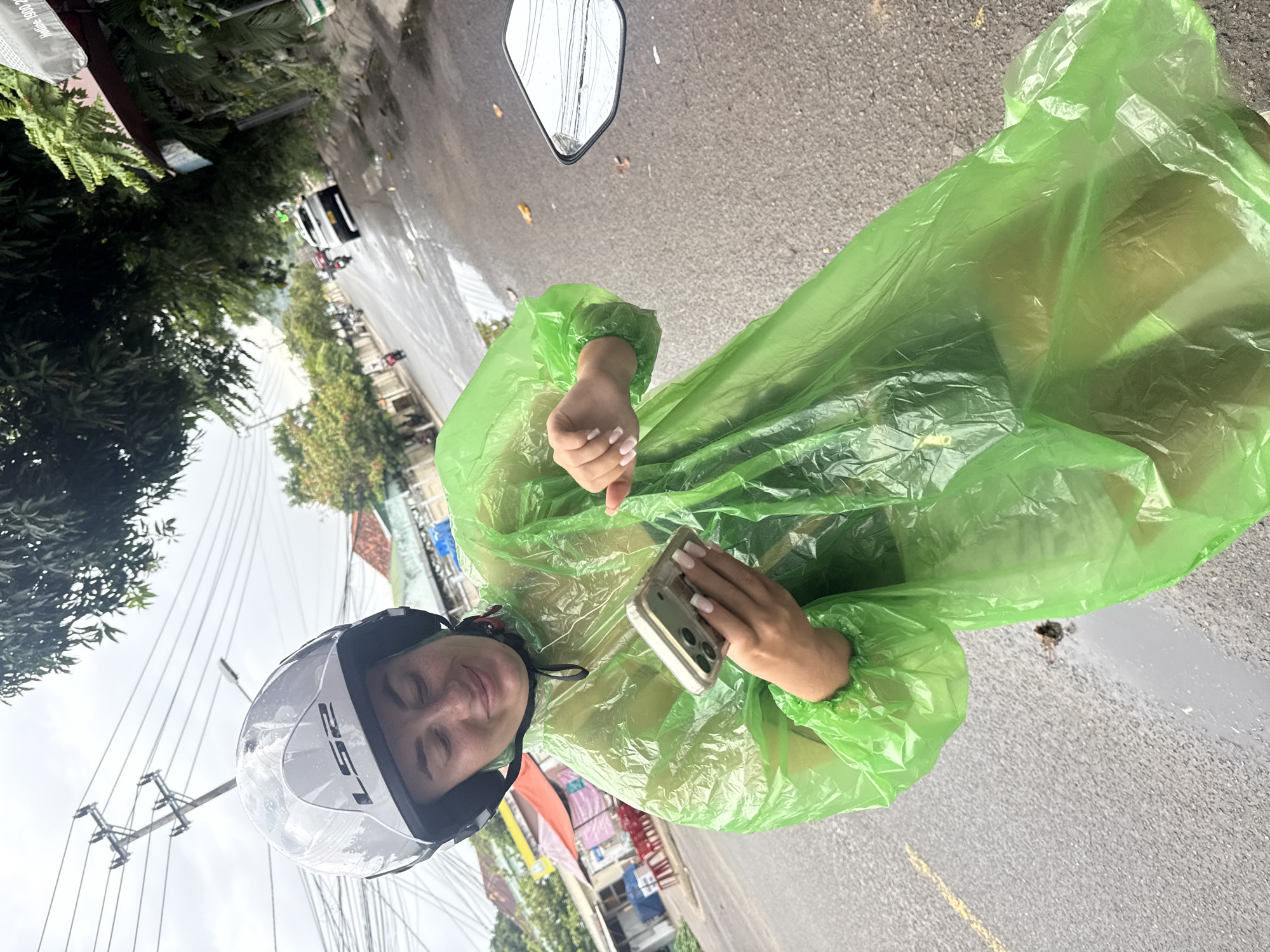 In Nha Trang, Vietnam, a woman wearing a bright green hooded poncho and a white helmet is looking down at her phone while holding it in her left hand. Her right hand is outstretched, and she is looking at her phone with a slight smile. The poncho is shiny and translucent, with some creases and folds visible. The woman has light-colored hair and her nails are painted a light pink. She is on a street that appears wet from rain, with puddles on the asphalt. In the background, there are trees, power lines, buildings with red-tiled roofs, and other vehicles, including a white car and several motorcycles. The weather appears overcast.