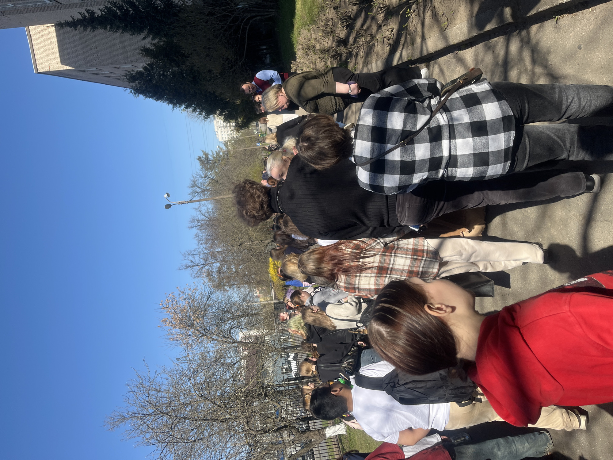 The image displays an outdoor scene under a clear, bright blue sky, indicative of a sunny day, likely mid-morning or afternoon given the strong sunlight and shadows. A diverse group of people, appearing to be mostly adults, are gathered in what seems to be a queue or waiting line. Many are viewed from behind or in profile, dressed in casual attire such as jackets, plaid shirts, hoodies, and jeans. Some individuals are standing, while others are seated on the paved ground, suggesting a period of waiting.
The setting is an urban environment, possibly a park pathway or a sidewalk adjacent to a street. The ground is a mix of concrete or asphalt, with some patches of grass and bare earth. Deciduous trees, some still largely bare-branched and others showing early buds or sparse foliage, line the area. In the background, a large, multi-story brick building is visible on the left, and other distant urban structures can be seen. A street light pole also stands in the mid-ground. The overall mood appears calm, with no discernible expressions of urgency or strong emotion among the individuals, implying a patient wait. No visible text is apparent on any objects or clothing. This scene is situated in Saint Petersburg, Russia, and appears to capture an early spring day.