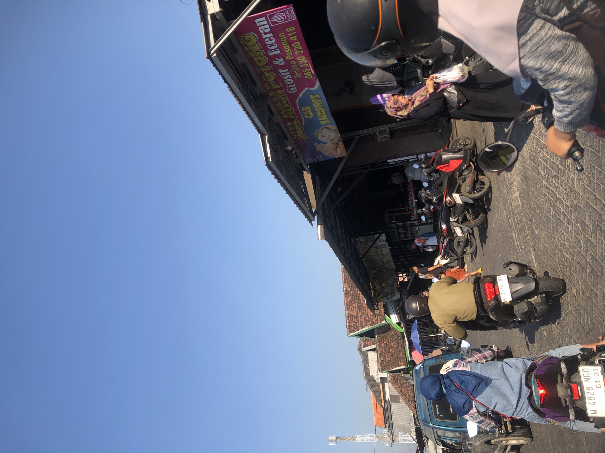 This is a street-level, eye-level, outdoor, daytime photograph taken in Kludan, Indonesia. The scene depicts a busy street with several motorcycles and a blue SUV. In the foreground, a person wearing a blue headscarf and a plaid shirt rides a motorcycle. Behind them, another person in a green jacket rides a motorcycle. Further down the street, a woman in a black abaya and a floral hijab is walking. A banner above a storefront reads "JUAL AYAM POTONG" and advertises "Grosir & Eceran," indicating a wholesale and retail chicken seller. The buildings have traditional tiled roofs, and a cell tower is visible in the distance. The ground is paved with cobblestones. The sky is clear and blue.
