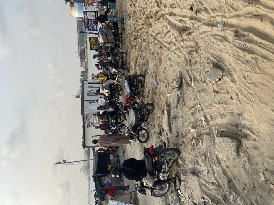 The scene displays a row of motorcycles parked in the sand near a body of water. People stand and gather around the bikes, while a man is seated on one, presumably about to ride off. Tire tracks crisscross the sandy terrain. Buildings and commercial signs are visible in the background. The sky appears overcast.