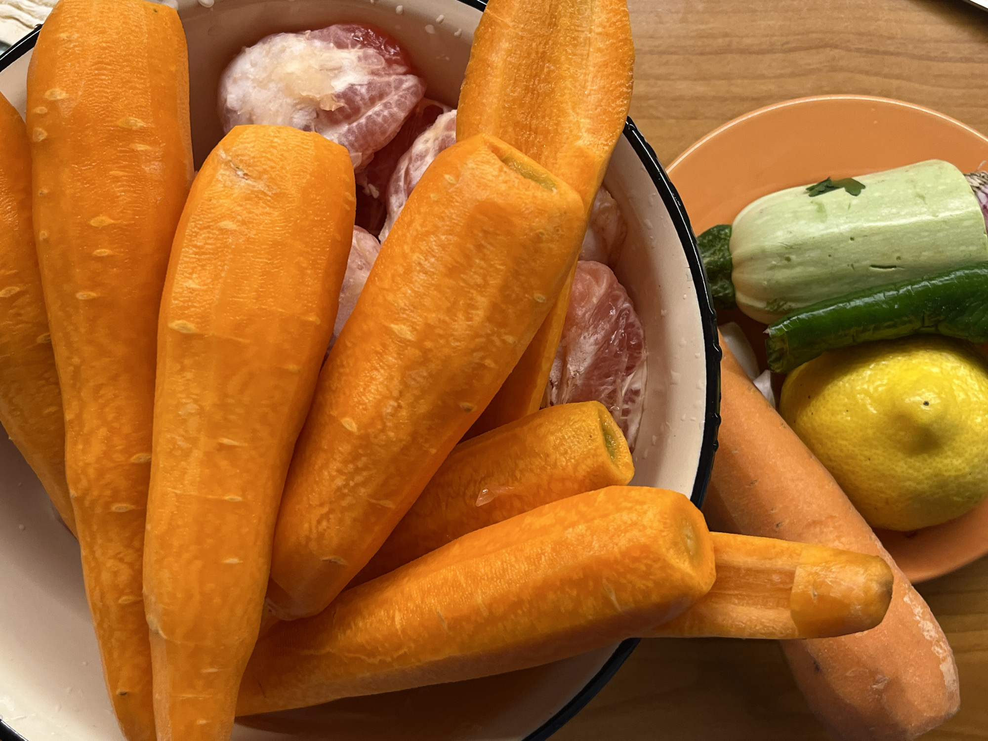 A close-up overhead shot shows peeled carrots and other vegetables arranged on a wooden surface. The carrots, a vibrant orange, are the most prominent subjects, piled in a white bowl. Some of the carrots are cut, revealing their green cores. Beneath the carrots, a few pieces of what appear to be peeled citrus fruit, possibly grapefruit, are visible. To the right of the bowl, a bright orange plate holds a lemon, a green bell pepper, and a light green zucchini. The lighting is natural, casting soft shadows and highlighting the textures of the produce. The scene is clean and appears to be set in a kitchen or dining area, though no specific details of the location are discernable beyond the wooden table.