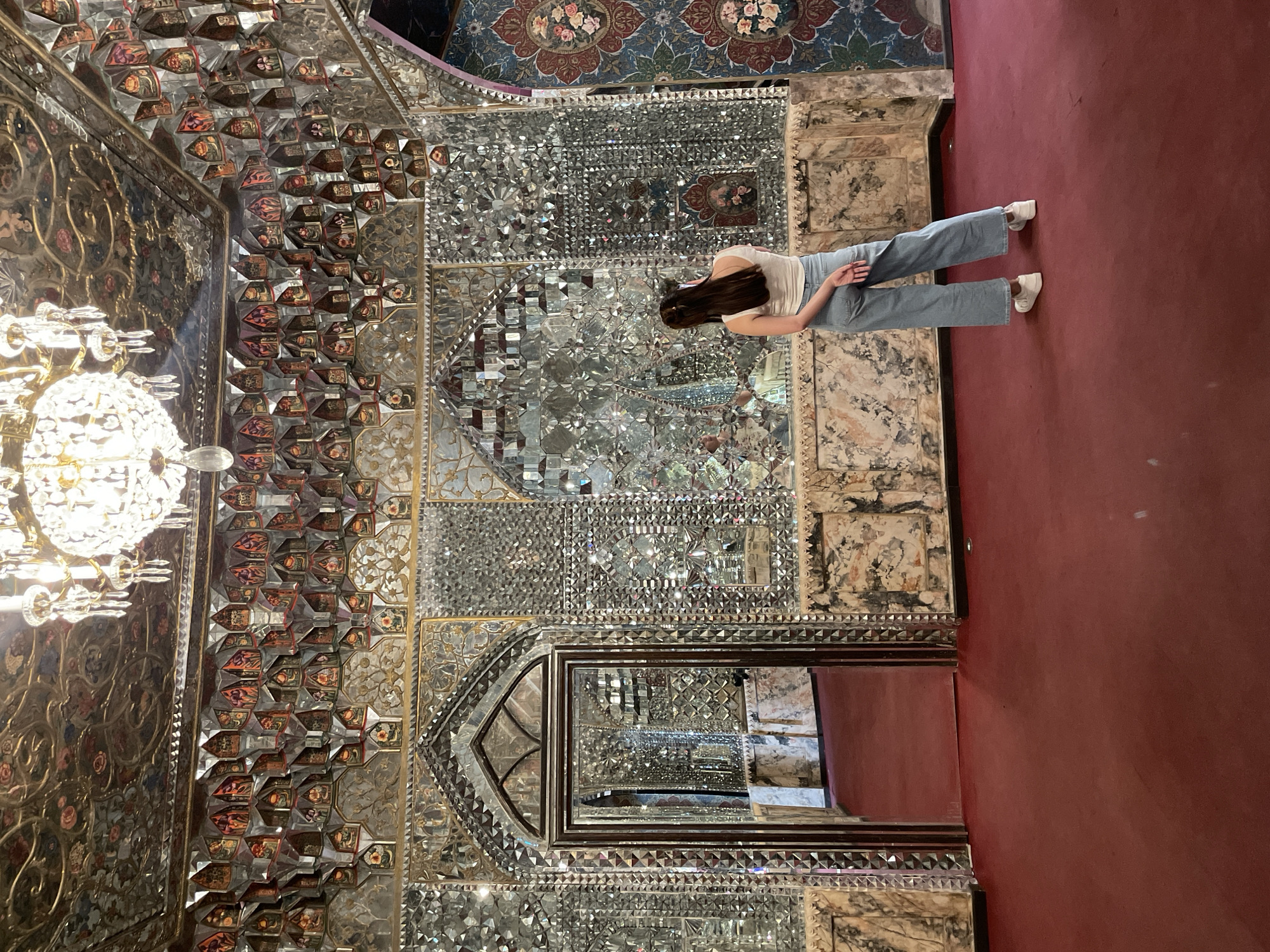 The image displays the interior of a lavishly decorated room, characterized by extensive and intricate mirror mosaic work (Shishehkari) covering the walls and ceiling. A person is central to the frame, viewed from the back, standing on a vibrant red carpet or rug.
The individual is a woman with long, dark hair, wearing a white sleeveless top, light blue wide-leg jeans, and white sneakers. She appears to be observing the room's ornate details.
The walls and ceiling are adorned with countless small, multifaceted mirror fragments arranged in elaborate geometric and floral patterns, creating a dazzling, kaleidoscopic effect. Interspersed within the mirrored surfaces are sections featuring detailed painted panels, possibly depicting flowers or other decorative motifs. A prominent, multi-tiered crystal chandelier hangs from the ceiling, its reflections adding to the room's brilliant shimmer. A large, dark-framed mirror, possibly serving as a doorway or an alcove, is visible towards the bottom right of the primary mirrored wall, reflecting other parts of the room. The luxurious and intricate design suggests a historical palace, museum, or an important cultural building, consistent with the location context of Tbilisi, Georgia, where such Persian-influenced architectural styles can be found. There is no visible text in the image.