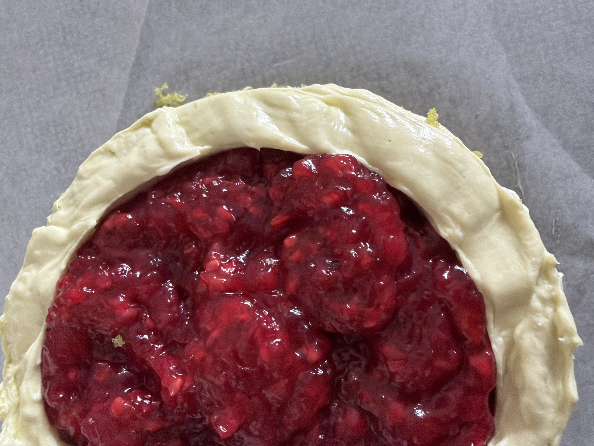 The image shows a close-up of a partially assembled raspberry galette on parchment paper. The filling is a glossy, dark red raspberry mixture. A thick, uncooked pastry crust surrounds the filling, folded in a decorative fashion. There are specks of green on the pastry, likely zest. The background is a plain gray parchment paper. The image provides no evidence of it being taken in Tel-Aviv, Israel, or of any people, subjects, or interactions.