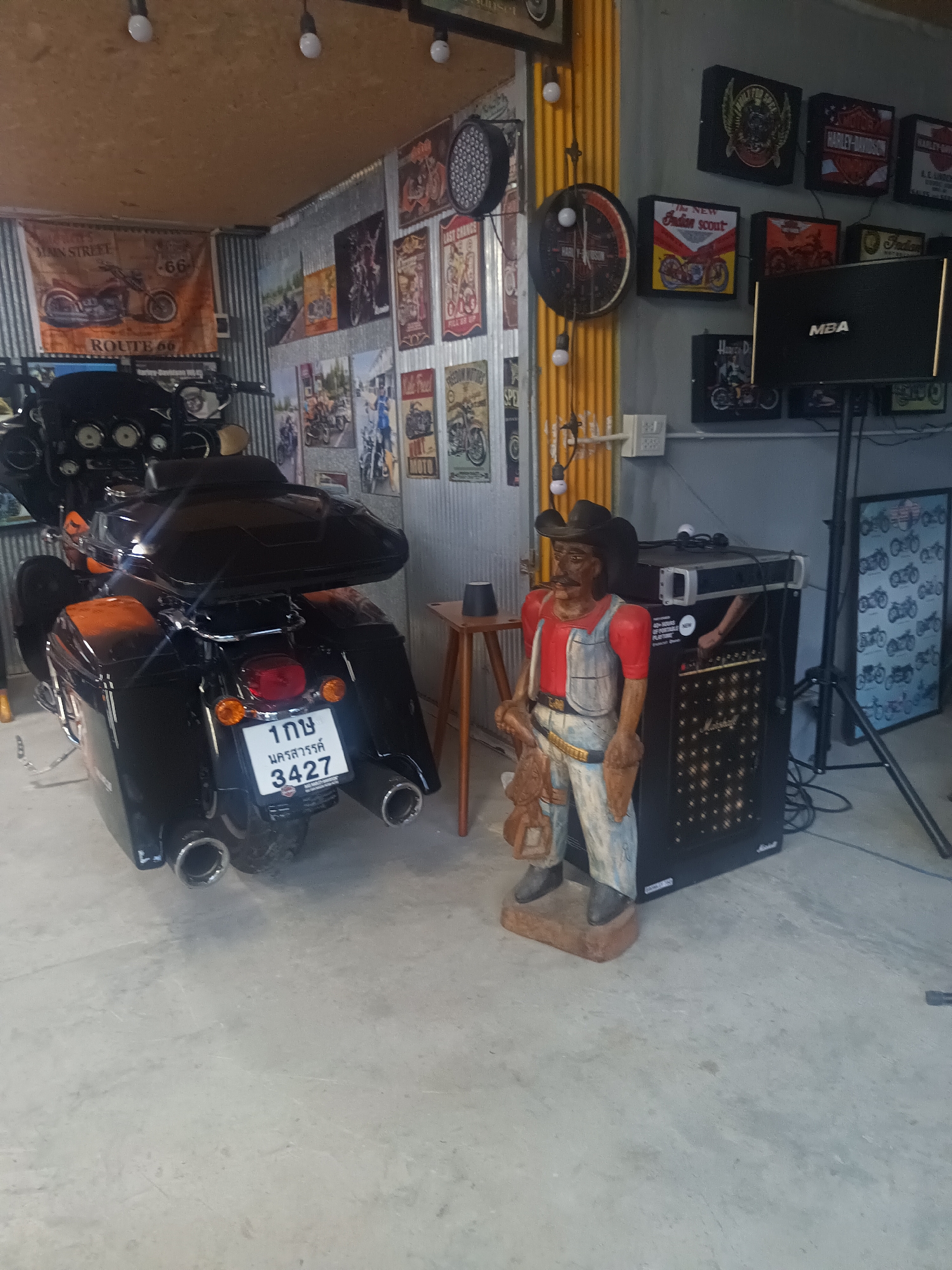 This image was taken indoors in the city of Surat Thani City Municipality, Thailand. The photograph features a black motorcycle in the foreground. To the right of the motorcycle is a cowboy statue dressed in overalls, a red shirt, and a hat. Behind the cowboy statue is a black amplifier and a speaker on a stand. The walls in the background are decorated with various motorcycle-themed posters and memorabilia. The environment appears to be a garage or showroom.