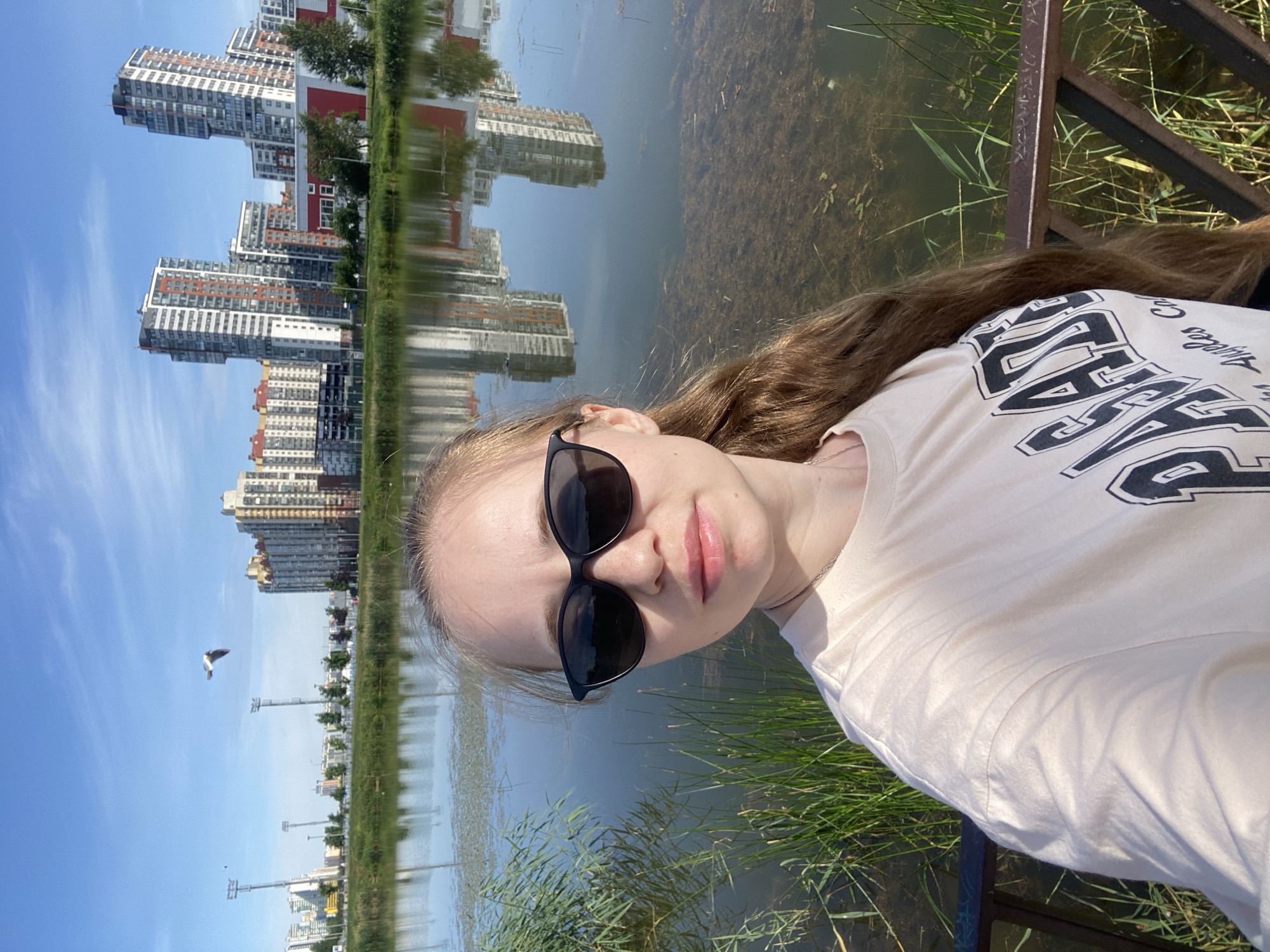 A young woman with long, light brown hair and sunglasses takes a selfie outdoors. She is wearing a light-colored t-shirt with a graphic print. The background shows a body of water with reflections of modern high-rise buildings. There are tall reeds and grass along the water's edge, and a metal railing in the foreground. In the sky, a bird is in flight. The architecture of the buildings and the presence of tall lampposts suggest an urban environment. This scene is consistent with the Novoorlovsky Park area in Saint Petersburg, Russia, known for its modern residential complexes and proximity to green spaces and water bodies.