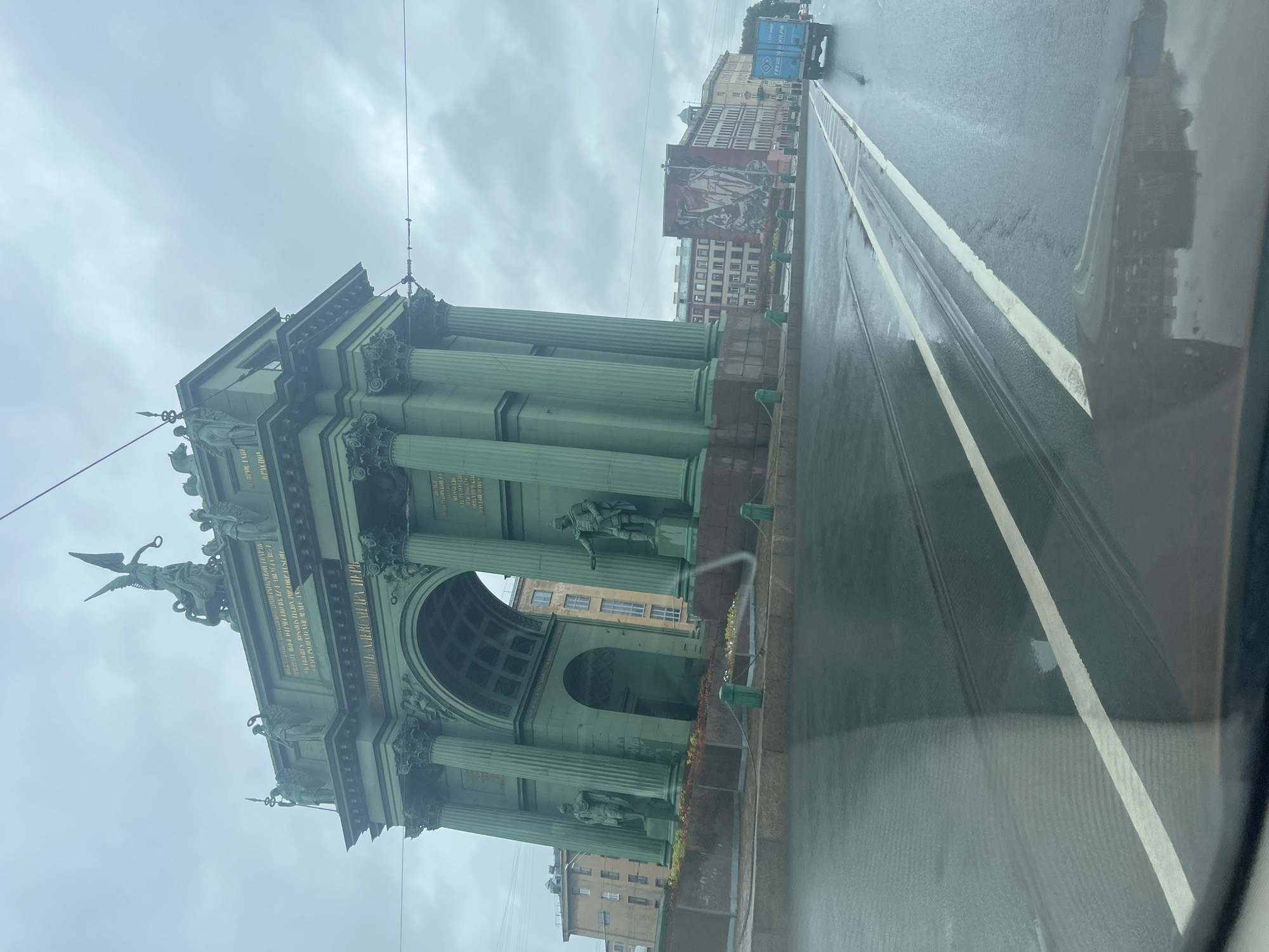 The image captures a view from a vehicle on a wet street in Saint Petersburg, Russia. The dominant structure in the scene is the Narva Triumphal Arch, a grand green archway adorned with statues and inscriptions. The arch is set against a cloudy sky. The wet road in the foreground reflects the arch and surrounding buildings. A blue truck is visible driving on the road. The architecture suggests a historical urban setting.
