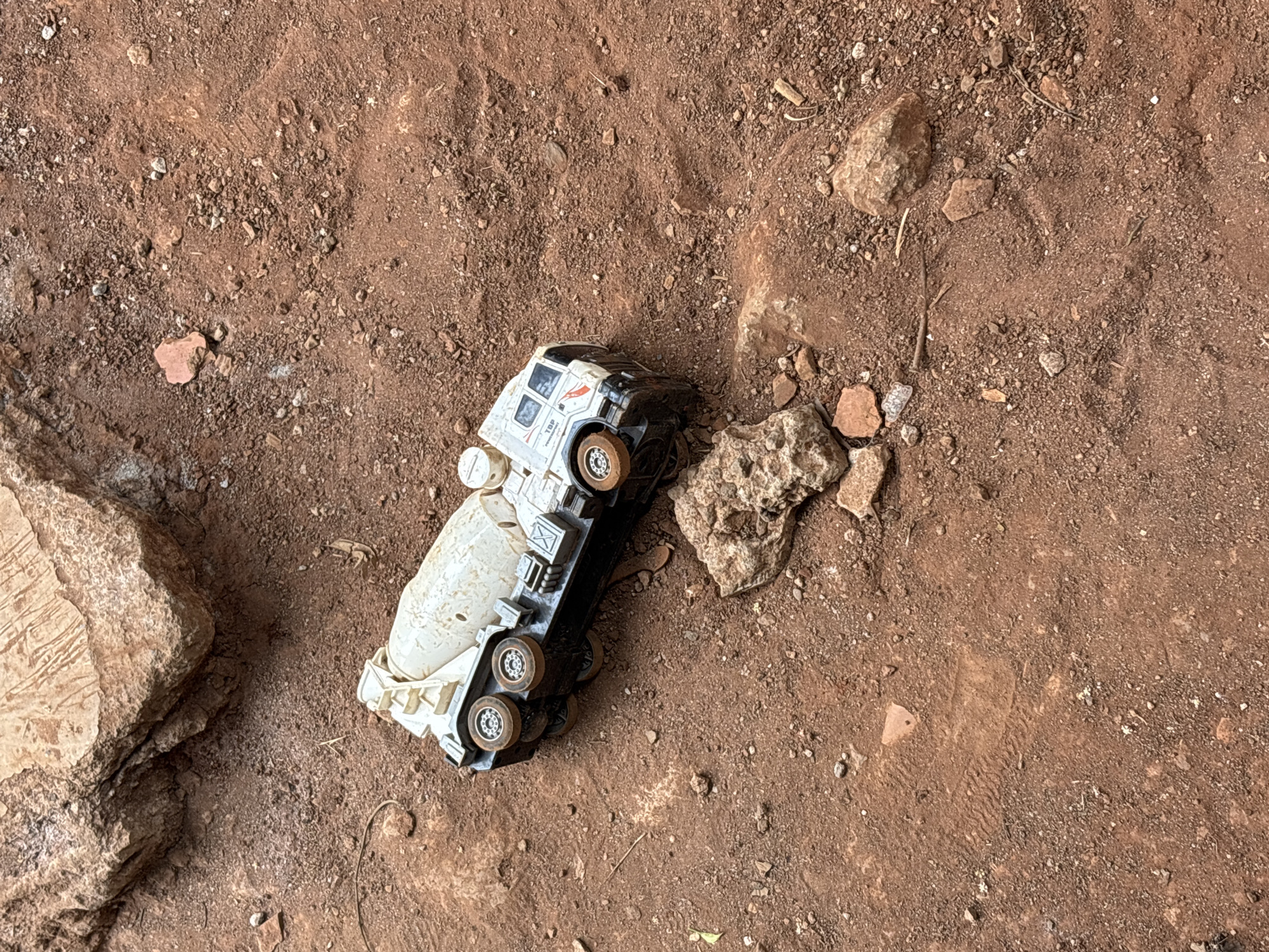 A close-up, high-angle shot shows a white and black toy cement mixer truck lying on its side in reddish-brown dirt and gravel. The truck is slightly dirty and appears to be overturned. To the upper left of the truck, a large, rough, tan-colored rock is partially visible. The ground is uneven, with smaller pebbles and some dried grass scattered around. The scene is outdoors, with natural light illuminating the objects. There are no people or other animals present in the image. The location is identified as Taman Rahayu, Indonesia, suggesting a residential or possibly a construction area.