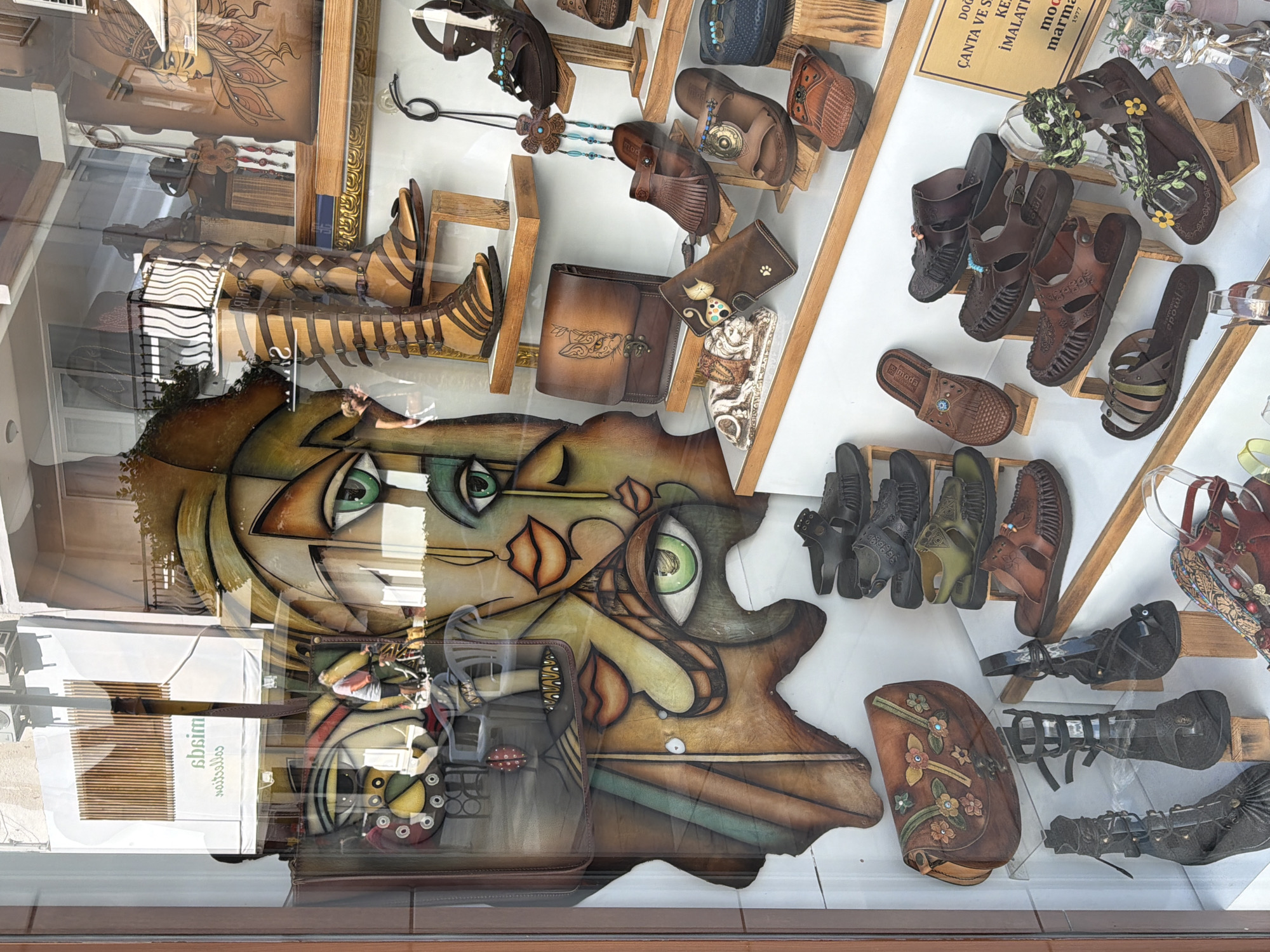 The image shows a shop window in Marmaris, Turkey, filled with various handmade leather goods, primarily women's shoes and handbags. The window display is arranged on wooden shelves. Prominently featured are several pairs of sandals and high-heeled shoes, many with intricate straps and embellishments. Handbags of different sizes and designs, including one with a cat embossed on it, are also visible. A large, abstract, colorful painting of faces is the backdrop behind some of the merchandise. Reflections of the street and a person riding a bicycle are visible in the window glass. A sign with Turkish text, including "MARMARIS," is visible on the right side of the display. The overall impression is of a boutique selling artisanal leather products.