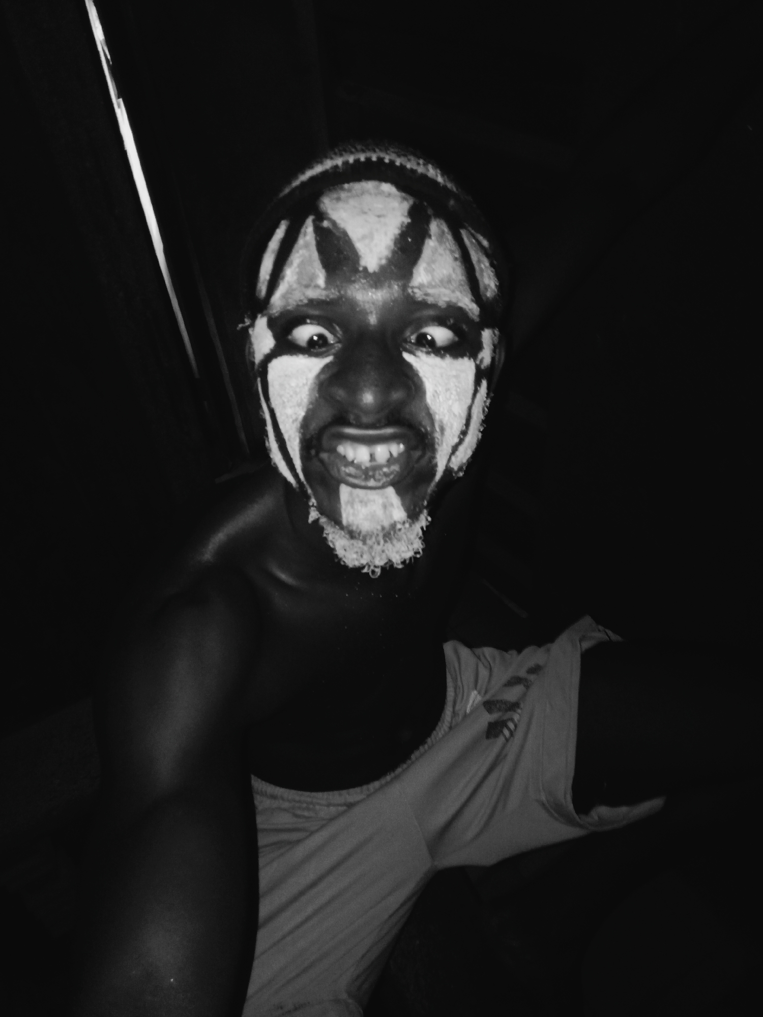 A monochrome, close-up portrait captures a person with elaborate white face paint against a dark background. The subject, with dark skin, wears stark white paint in bold patterns, including a prominent "X" or star-like design on the forehead, thick lines around the eyes, and a textured, beard-like application covering the chin and mouth.
The person's eyes are wide and intensely focused on the viewer, accompanied by a wide grimace revealing their teeth. This expression conveys an energetic, perhaps wild or theatrical, emotional state. A textured head covering is visible on the top of their head. A light-colored, possibly white or off-white, garment is draped over the person's left shoulder and arm, its fabric appearing soft and creased.
The image is dimly lit, with a strong, directional light source from the upper left casting highlights on the subject's left arm, shoulder, and the left side of their face. The background is almost entirely black, emphasizing the subject's painted features and expression. The overall atmosphere is dark and intimate, suggesting an indoor setting or nighttime. No visible text is present. The scene is set in Onitsha, Nigeria, implying a cultural or performance context for the face paint.