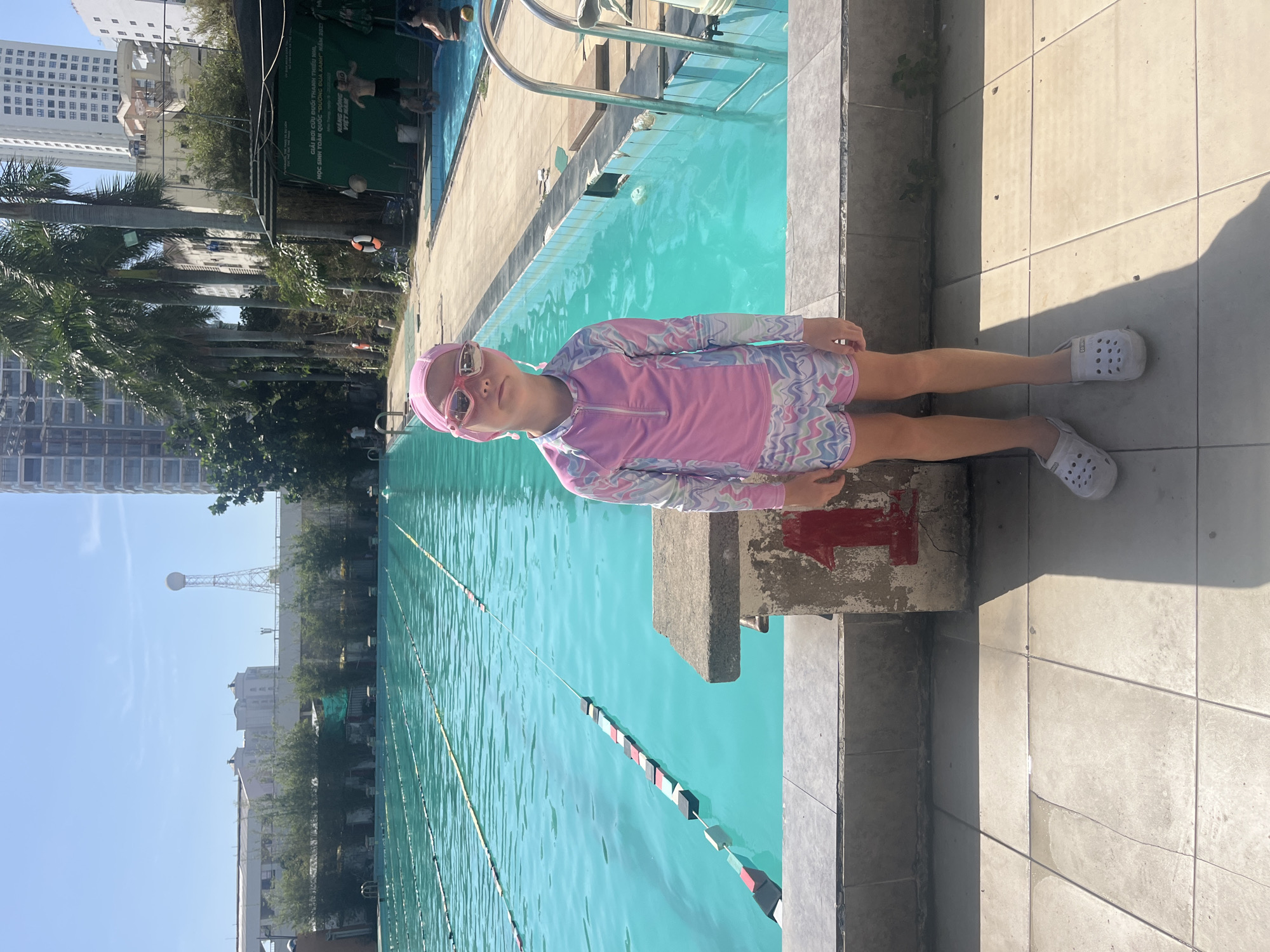 An outdoor swimming pool complex in Nha Trang City, Phước Hải Ward, Vietnam, is depicted under bright, clear daylight. A young Caucasian girl, approximately 6-8 years old, stands centered in the foreground, facing the camera with a neutral expression. She wears a long-sleeved pink and patterned swim shirt, matching patterned swim shorts, a pink swim cap, and pink-rimmed goggles. Her feet are clad in white Crocs-style sandals. She stands on a tiled concrete path next to a weathered concrete diving block, marked with a large red number "1".
To her left, a large, turquoise-blue lap pool extends into the background, equipped with multiple lane lines featuring red, black, white, and yellow buoys. On the far side of this pool, a row of low green bushes and buildings is visible, with a tall, white lattice tower topped by a sphere in the distance under a clear blue sky.
To the girl's right and behind her, separated by a concrete walkway, is a shallower, smaller swimming pool where several individuals are present. An adult male stands waist-deep, and another adult and a child are partially visible in the water near metal pool steps. A red and white lifebuoy hangs on a pole near this smaller pool. Tall palm trees line the area between the pools and the background, where modern high-rise buildings are visible. A green banner with white Vietnamese text, advertising a "Youth Swimming and Drowning Rescue Competition" held in Nha Trang in May 2023, is prominently displayed on the right side. The ground is paved with light grey square tiles, and strong shadows indicate the sun is high in the sky.
