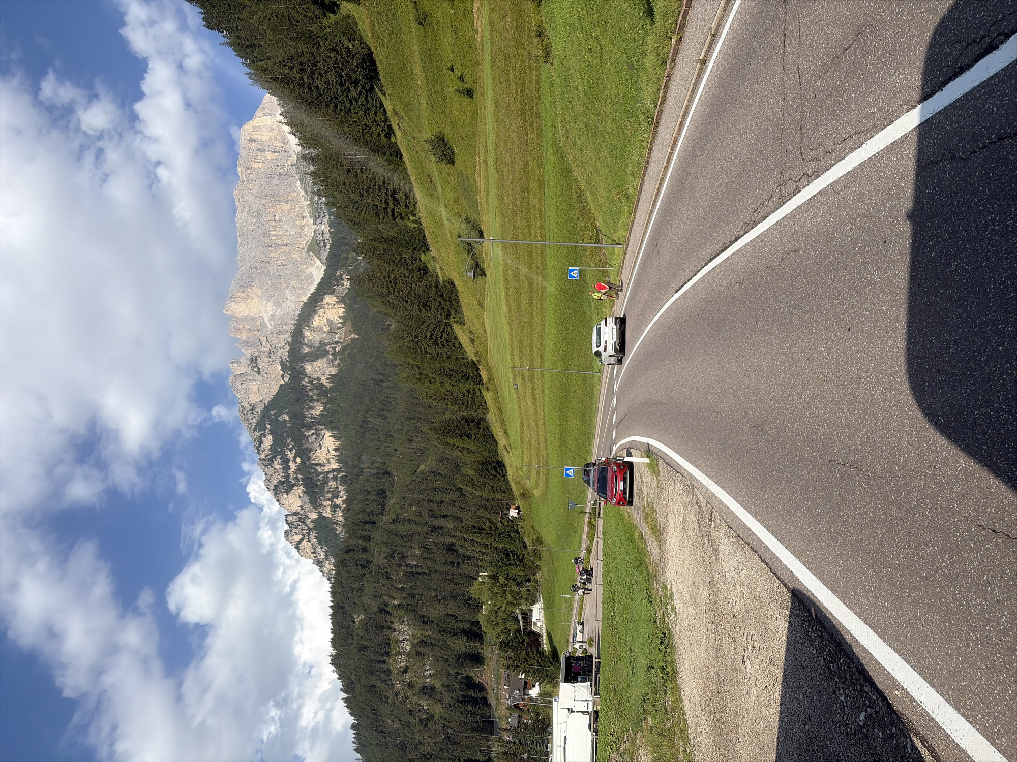The image captures a scenic mountain road in the vicinity of Mazzin, Italy. A paved road with white lane markings curves through a landscape dominated by lush green hills and a dense pine forest that climbs the mountainside. In the distance, a majestic, rocky mountain peak with sheer cliffs stands against a backdrop of a partly cloudy blue sky.
In the foreground, the road is traveled by a white car in the right lane and a red car parked on the left side of the road, near a gravel shoulder. Further down the road, a pedestrian crossing sign is visible. To the left of the road, a parking area can be seen with several motorcycles and a white trailer. Two individuals, one wearing a red shirt and the other in a yellow vest, are standing near the white car. The environment suggests a summer day with pleasant weather, ideal for outdoor activities in the Dolomites region.