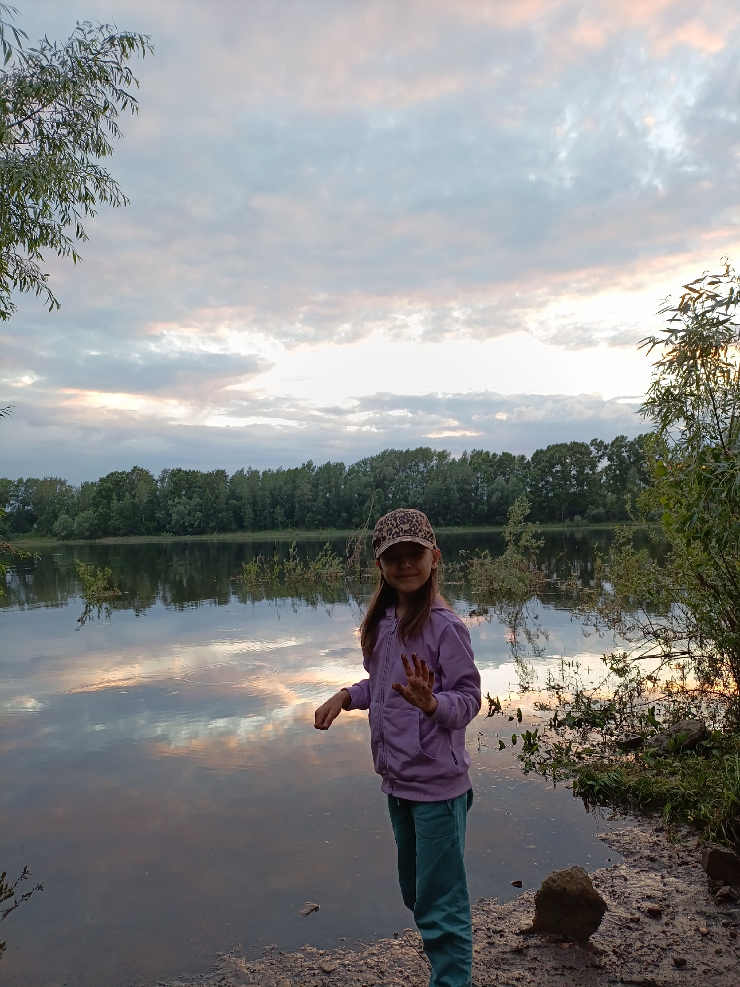 An outdoor, eye-level, medium shot captures a young girl standing on the muddy bank of a wide river. The girl, wearing a purple hooded jacket and teal pants, has her hands raised, palms facing outward, and appears to be looking at the viewer. She also wears a leopard-print baseball cap. The river water reflects the cloudy, pastel-colored sky and the dense line of green trees on the opposite bank. Vegetation grows along the immediate shoreline, and a few rocks are scattered on the ground. The location is the Kama River in the city of Chaikovsky, Russia.
