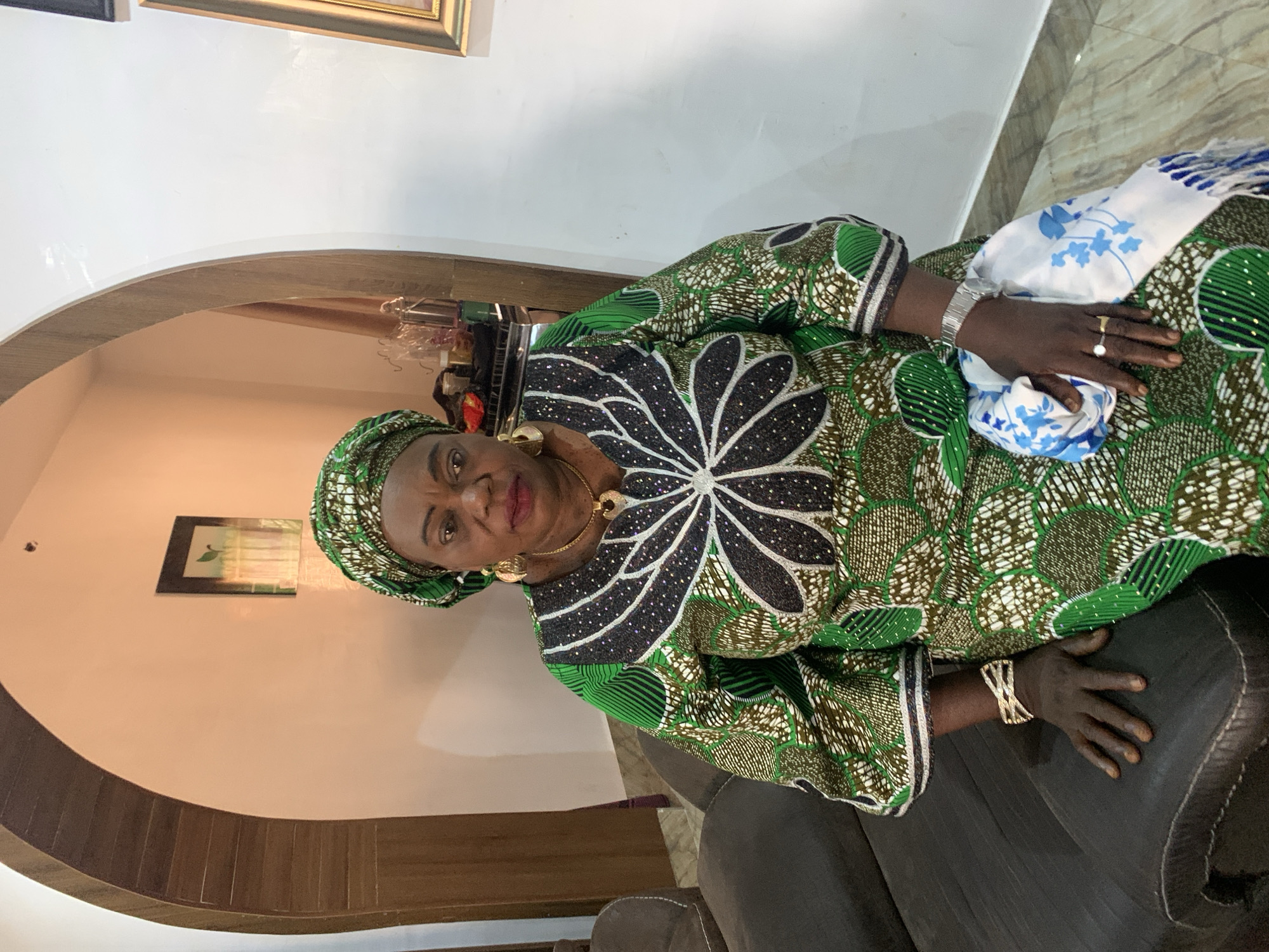 The image shows a mature woman with brown skin, wearing a green and brown patterned dress with a large, stylized black and white flower design on the front. She has a matching headwrap and gold jewelry, including earrings and a necklace. She is sitting on a dark, possibly leather, sofa. She wears a silver watch on her left wrist and a gold ring with a pearl on her right hand, which is resting on a white scarf with blue accents. The background shows an interior space with a curved wooden architectural detail, light-colored walls, and a framed picture on the wall. The location is identified as Jos, Nigeria.
