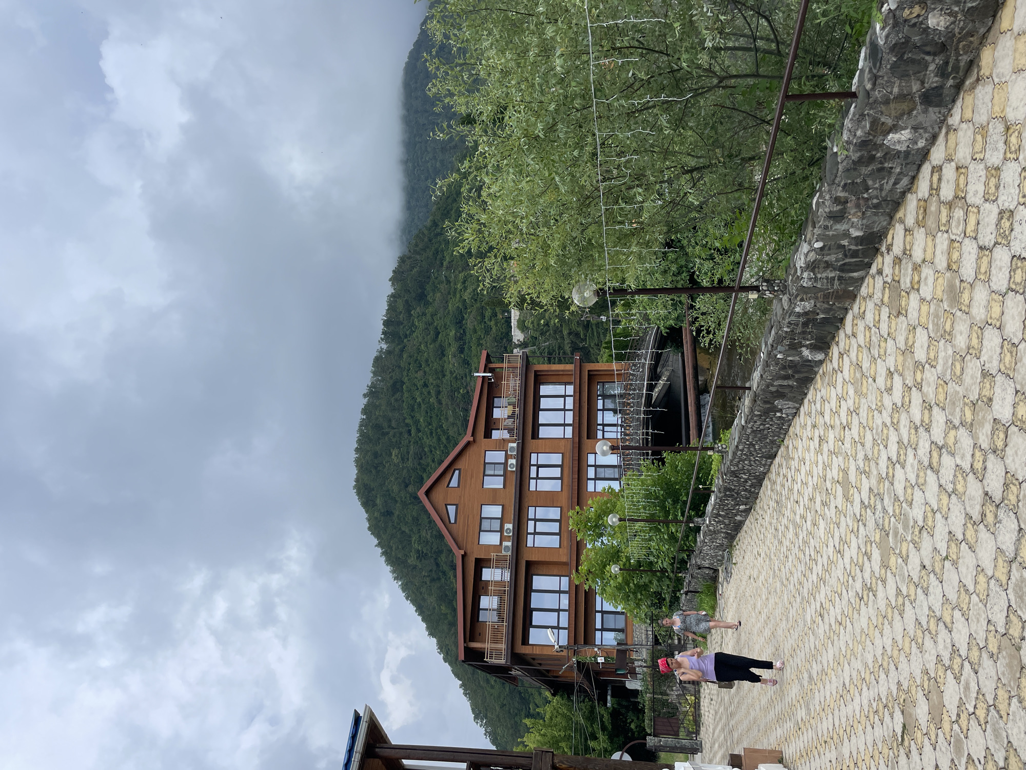 In the city of Abaata, Abkhazia, a hotel with a wooden facade and multiple stories is situated against a lush green mountainside partially obscured by clouds. Two women are walking on a paved pathway in front of the hotel. The pathway is made of interlocking hexagonal and square tiles in shades of yellow and grey. A stone wall borders the pathway on the right, with a metal railing on top. Trees and foliage surround the hotel and the pathway, with some trees bearing string lights. A bridge is visible behind the hotel, spanning a body of water. The sky is overcast with heavy clouds.