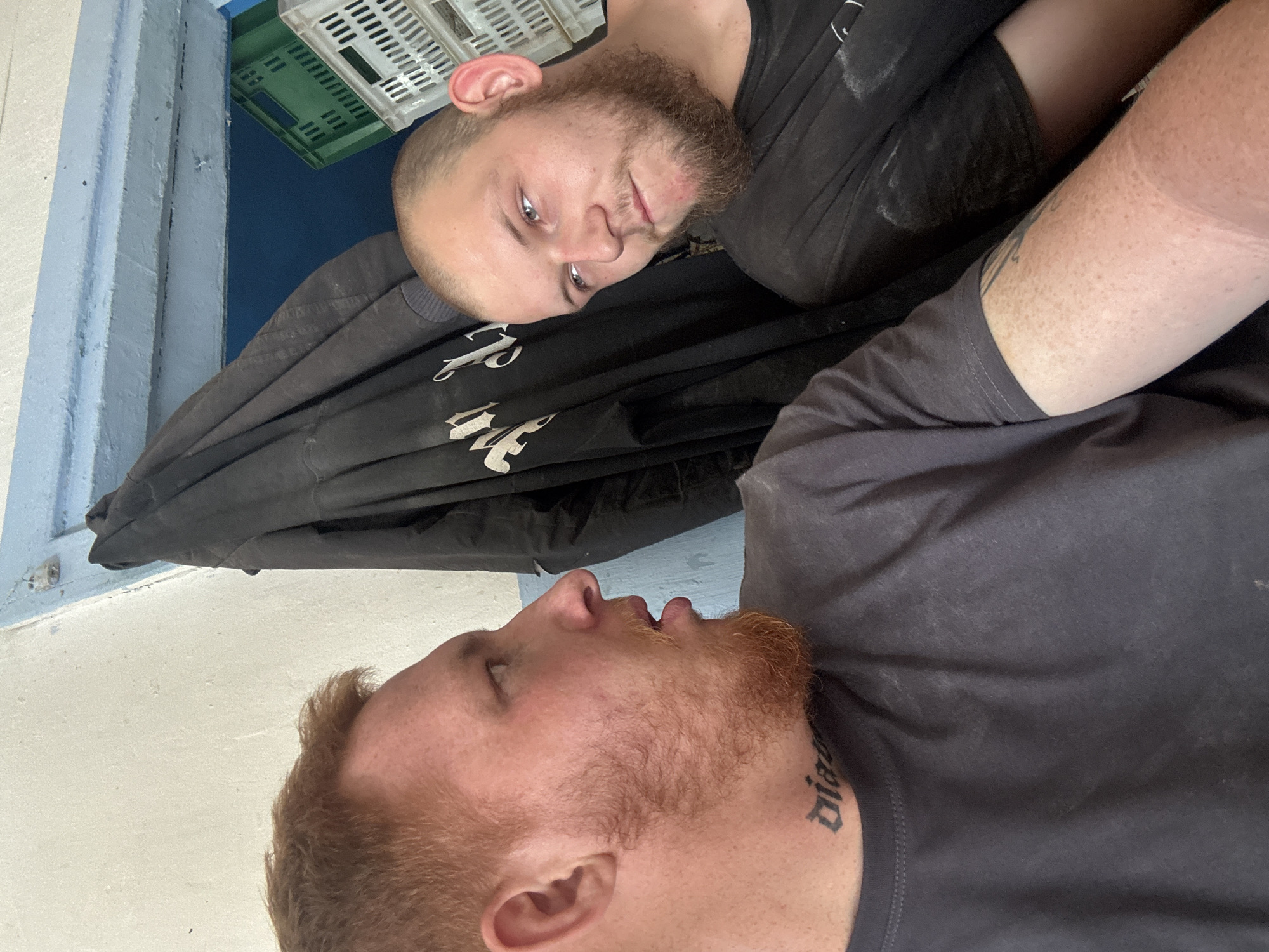 Two men are captured in a close-up, profile shot, appearing to be in a dimly lit, possibly industrial or storage area. The man on the left has ginger hair and a beard, with a tattoo on his neck that reads "diana." He is looking off to the right. The man on the right has short brown hair and a beard, and is looking down and to his left. He has a tattoo on his arm. They are both wearing dark t-shirts. In the background, a black jacket is hanging, and behind that, blue walls and white and green plastic crates are visible. The lighting suggests it could be daytime, but the indoor setting makes it difficult to ascertain the exact time or location.