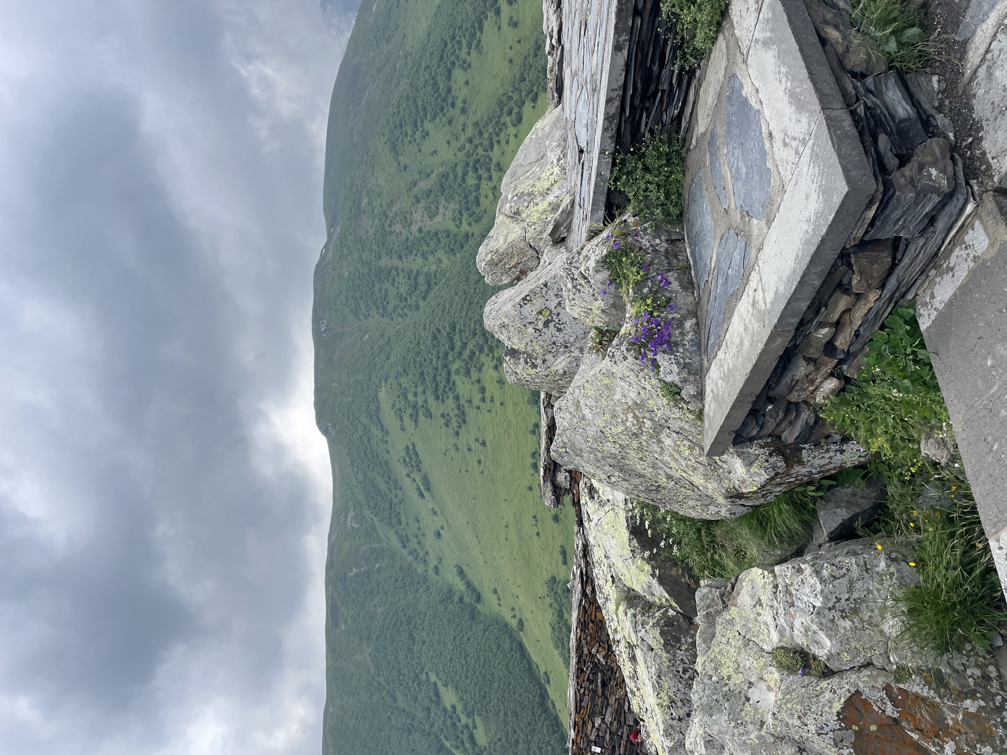 The image shows a mountainous landscape with a cloudy sky. In the foreground, there are large rocks and stone structures. A person wearing a red hat is visible to the far left, partially obscured by rocks. The background features a vast, green, tree-covered mountain slope. This scene is likely from Stepantsminda, Georgia, known for its dramatic mountain scenery and ancient architecture, such as Gergeti Trinity Church, which is often photographed from similar vantage points.