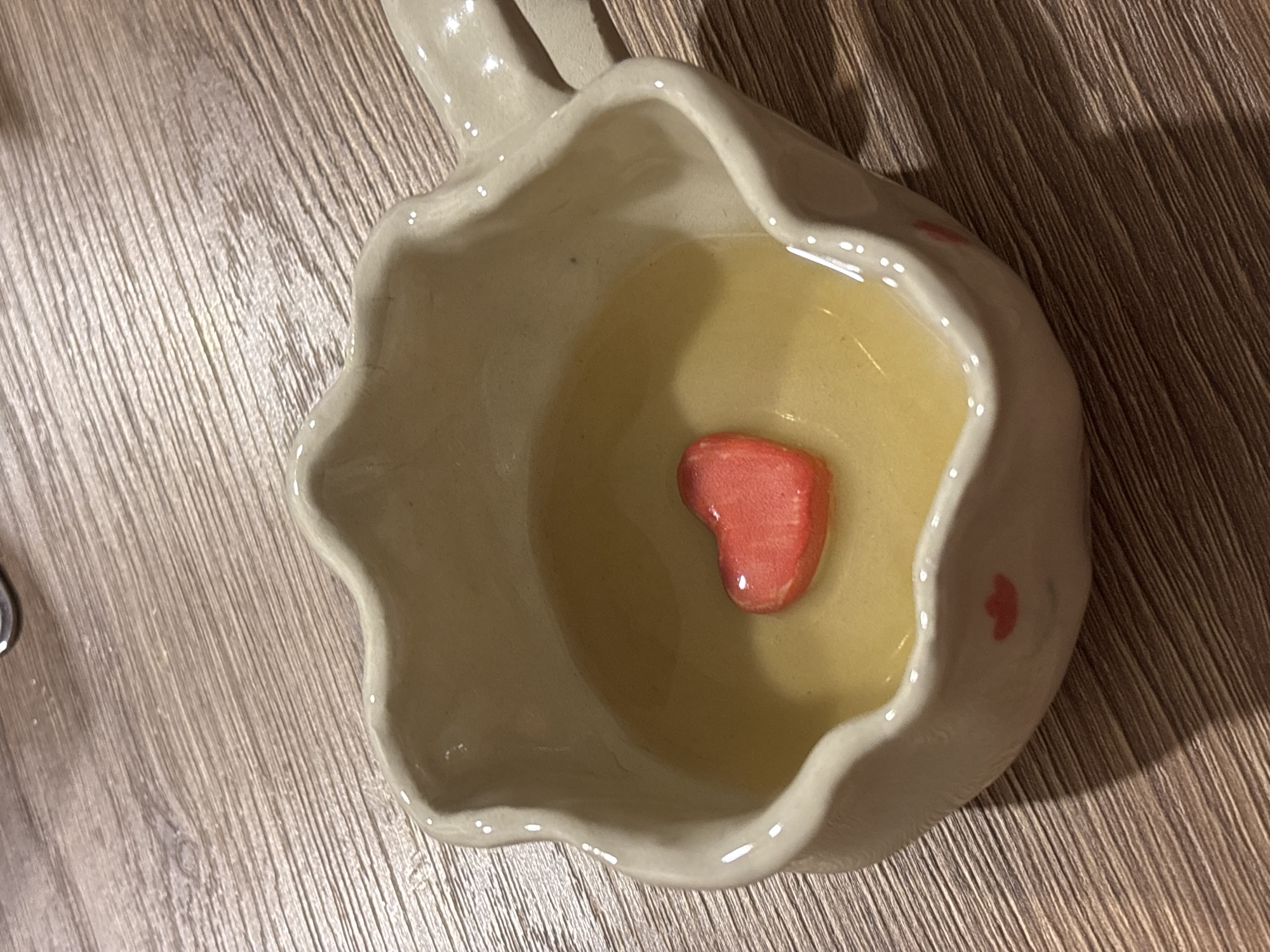 A ceramic mug with a scalloped edge sits on a wooden surface. The mug contains a light yellow liquid with a single red, heart-shaped gummy candy submerged in the center. The mug is off-white with red heart decorations on the exterior. The mug is oriented at an angle, with the handle partially visible in the upper left corner. The wooden surface has a light brown, striped pattern.