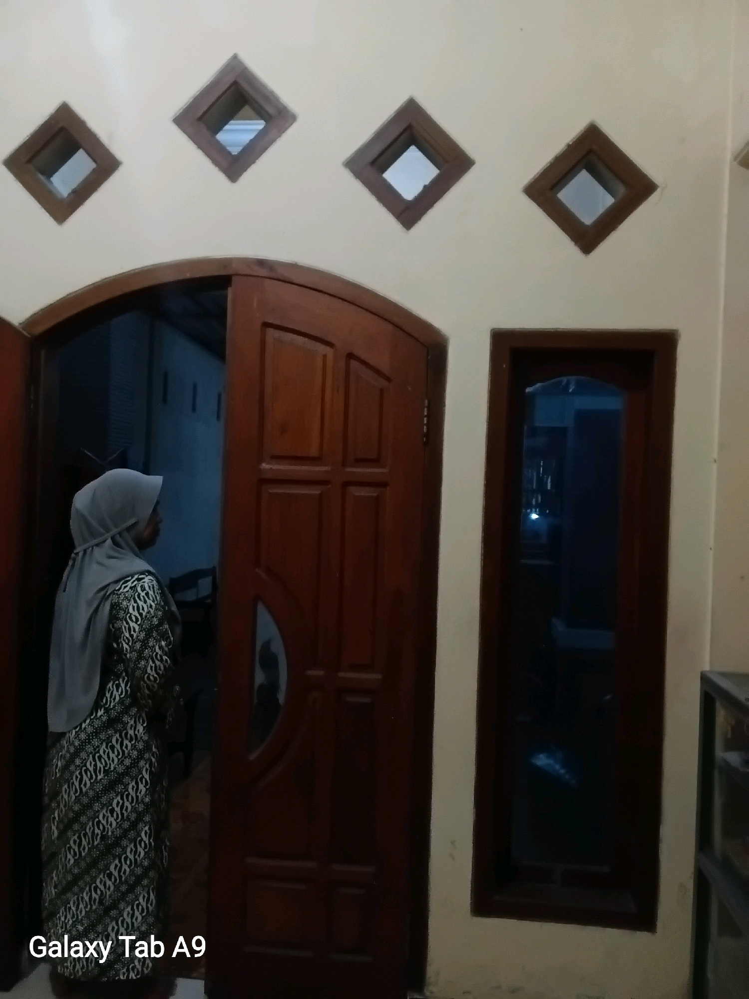A woman wearing a gray hijab and a patterned dress stands in an open doorway. The doorway is made of dark wood and has a curved top. To the left of the doorway is a tall, narrow window with dark wood trim. Above the doorway and window, on the cream-colored wall, are four diamond-shaped openings with dark wood frames. The image is watermarked with "Galaxy Tab A9" in the bottom left corner. The setting appears to be the exterior of a building, possibly a home, in Trenggalek, Indonesia. The lighting suggests it is evening or nighttime, with some interior lights visible through the doorway and window.