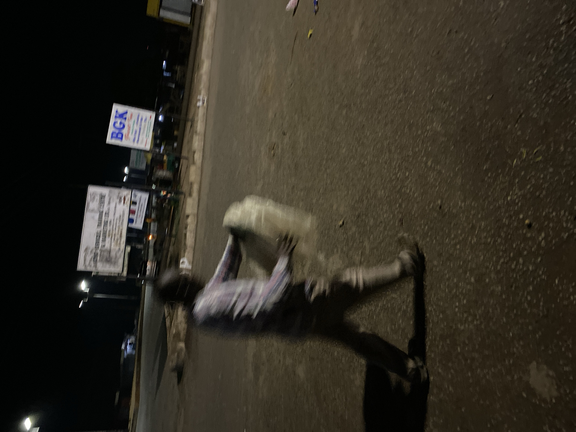 A blurry, night-time photograph captures a person walking across a paved road in Bukuru, Nigeria, carrying a large, white, translucent bundle. The individual is wearing a checkered long-sleeved shirt and light-colored trousers. The background shows streetlights and two billboards. One billboard, to the left, is partially obscured and displays text in English. The other billboard, to the right, prominently features the blue text "BGK". The road surface appears damp and reflects the ambient light, suggesting it may have recently rained. The overall atmosphere is that of a quiet, urban street at night.