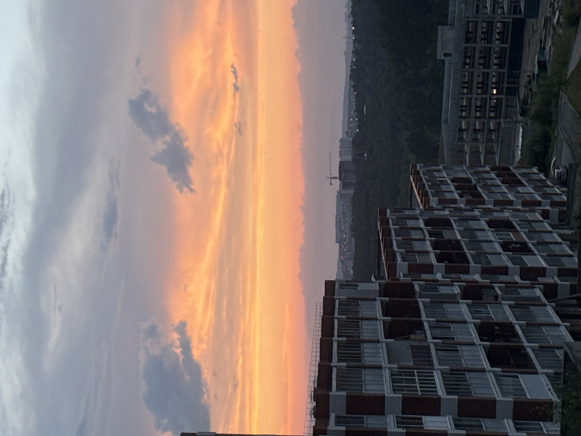 An outdoor, eye-level, medium shot captures a cityscape at dusk with a vibrant sunset. The sky is filled with shades of orange and pink, transitioning to grey clouds at the top. In the foreground, two multi-story buildings are under construction, with visible brickwork and concrete structures. The building on the left is partially completed, while the one on the right is a framework of floors and columns. Beyond these buildings, a dense forest stretches into the distance, dotted with more construction and city lights. The overall scene depicts urban development within a natural landscape during sunset.