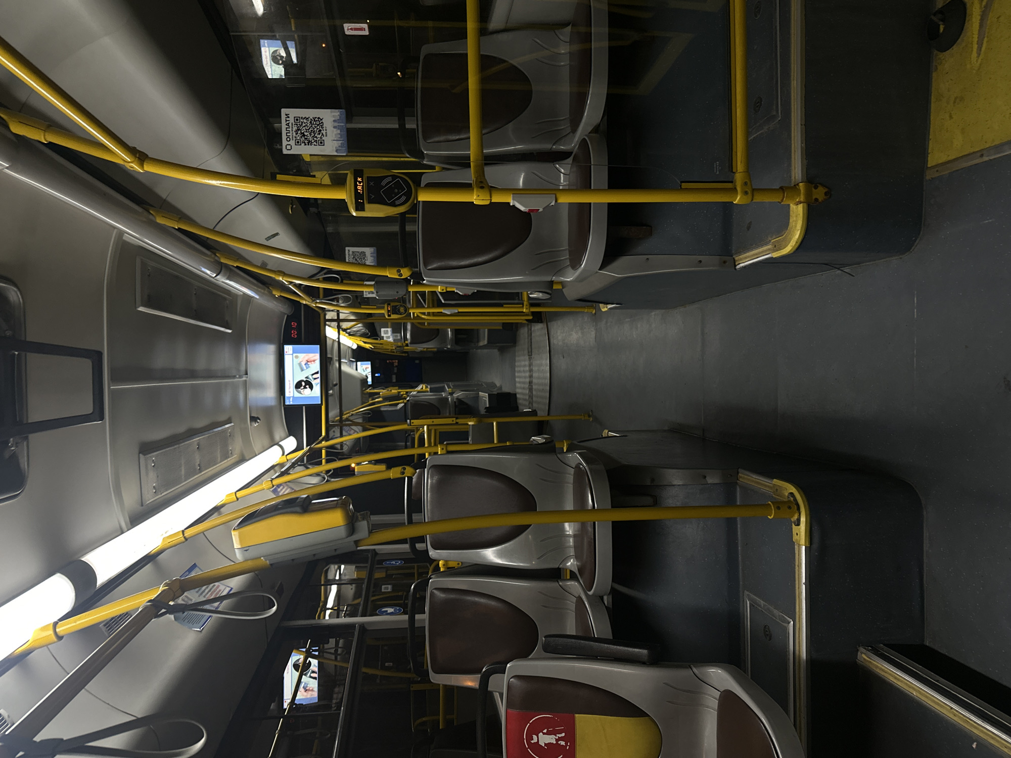 The image depicts the interior of a public transportation vehicle, likely a bus, in Minsk, Belarus. The scene is viewed from the perspective of someone standing in the aisle, looking towards the rear of the vehicle. The seats are arranged in pairs on either side of the aisle. They have a grey plastic shell with brown upholstered cushions. Yellow handrails are prominently featured throughout the interior, running along the ceiling and vertically between seats, providing support for passengers. The floor is dark grey with yellow accents.
Screens are visible mounted on the walls, displaying information or advertisements. One screen on the left shows a person's profile and the text "00:10". Another screen on the right displays a QR code for payment with the text "ОПЛАТИ" (meaning "Pay" in Russian). There are also warning signs with symbols, including one on the left seat showing a person with a cane, indicating priority seating.
The lighting is provided by long fluorescent tubes mounted on the ceiling, casting a cool, even light. The overall impression is that of an empty public transport vehicle, suggesting it is either not in service, or during a period of low passenger occupancy. The environment appears clean and well-maintained.