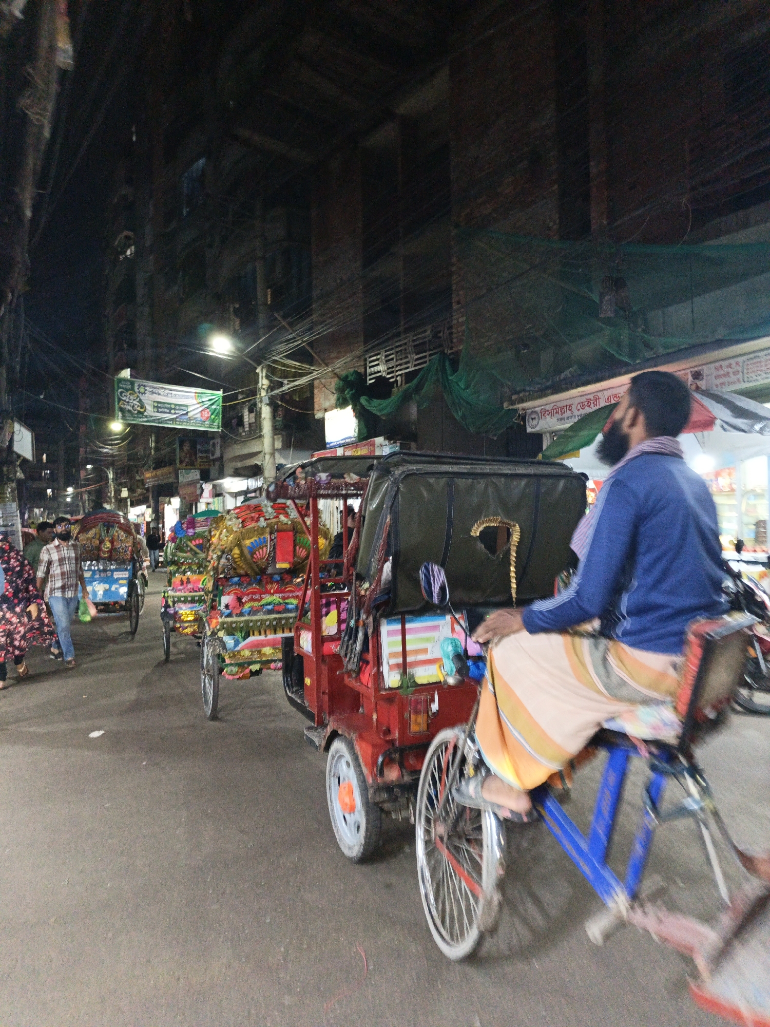 This nighttime image captures a bustling street scene in a dense urban environment, likely in South Asia. The primary activity involves transportation and pedestrian movement along a narrow, well-lit street.
In the foreground, a man with a beard, wearing a blue long-sleeved shirt and a striped *lungi* or similar lower garment, rides a blue-framed cycle-rickshaw, facing away from the viewer. His rickshaw has a black canopy and a decorative golden element resembling a question mark on the back. To the left, several elaborately decorated cycle-rickshaws, adorned with vibrant colors, patterns, and fringes, are visible, either stationary or moving. One prominent rickshaw features intricate designs in red, gold, blue, and green.
Other subjects include pedestrians on the left side of the street; a man in a plaid shirt and jeans walks alongside a woman in a dark, floral-patterned dress. Buildings with multiple stories line both sides of the street, their facades partially obscured by darkness and overhead structures. The street is illuminated by various artificial lights, including streetlights and shop lights, creating a bright central strip against the dark backdrop.
Notable details include a dense network of overhead electrical wires and cables crisscrossing the sky, characteristic of many urban areas in the region. Green safety netting or tarps are visible draped over some upper structures. A prominent green banner with white Bengali script hangs across the street. Another sign, partially visible in the background above the rickshaw rider, reads "বিসমিল্লাহ ডেইরী এন্ড" (Bismillah Dairy &) in Bengali script. The overall atmosphere is busy and active, reflecting everyday life after dark.