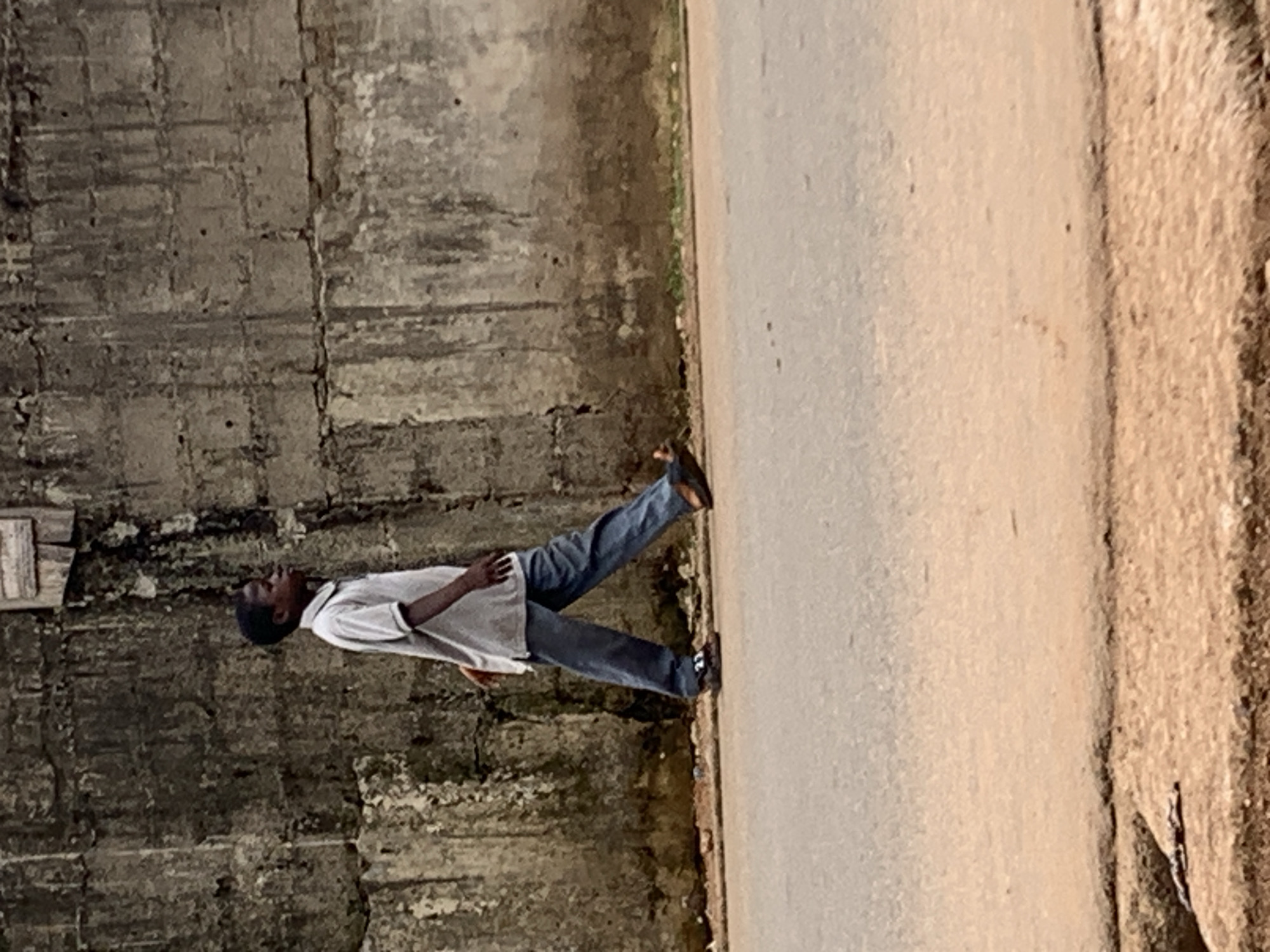 A man is walking alongside a concrete wall and road in Akwanga, Nigeria. The man is wearing a light-colored short-sleeved shirt and dark-colored jeans. He is looking upwards and walking on the side of a paved road next to a textured concrete wall. The background includes a road and some greenery. The wall appears weathered with visible cracks and signs of aging.