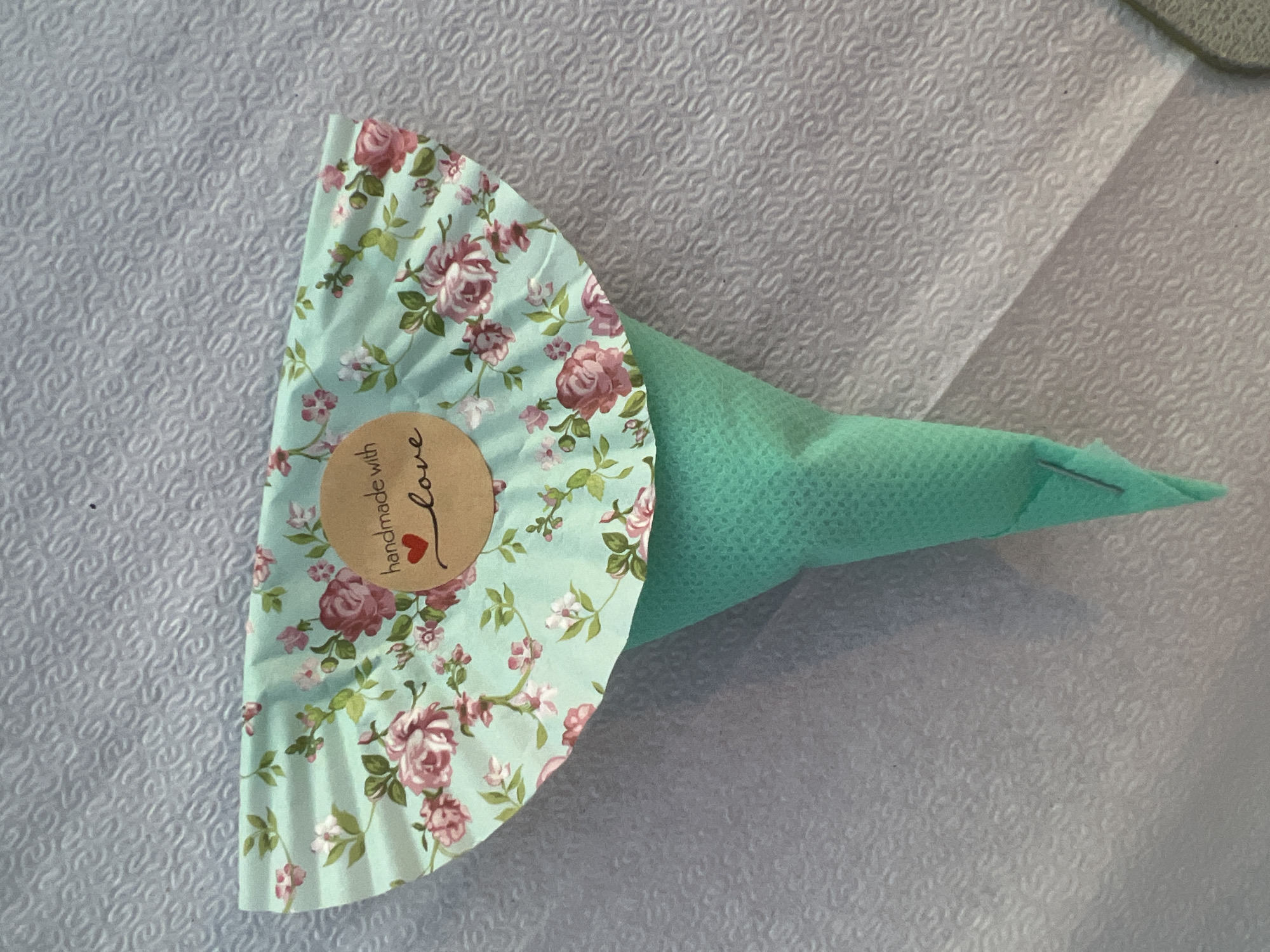 A close-up, top-down shot shows a handmade gift wrapped in a light turquoise paper with a floral pattern of pink roses and green leaves. A circular brown sticker with handwritten text "handmade with love" and a small red heart is attached to the paper. The gift is in the shape of a cone, made of a green textured material, and sits on a textured white surface that appears to be a tablecloth or placemat. There is a subtle shadow cast on the right side of the object. The specific location in Eretria Municipal Unit, Greece, cannot be determined from the visual information provided.